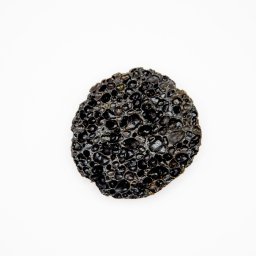 Justyna Tokarz - Czarny, porowaty kamień o nieregularnym kształcie na białym tle, przypominający pumeks lub skałę wulkaniczną z widocznymi pęcherzykami powietrza.