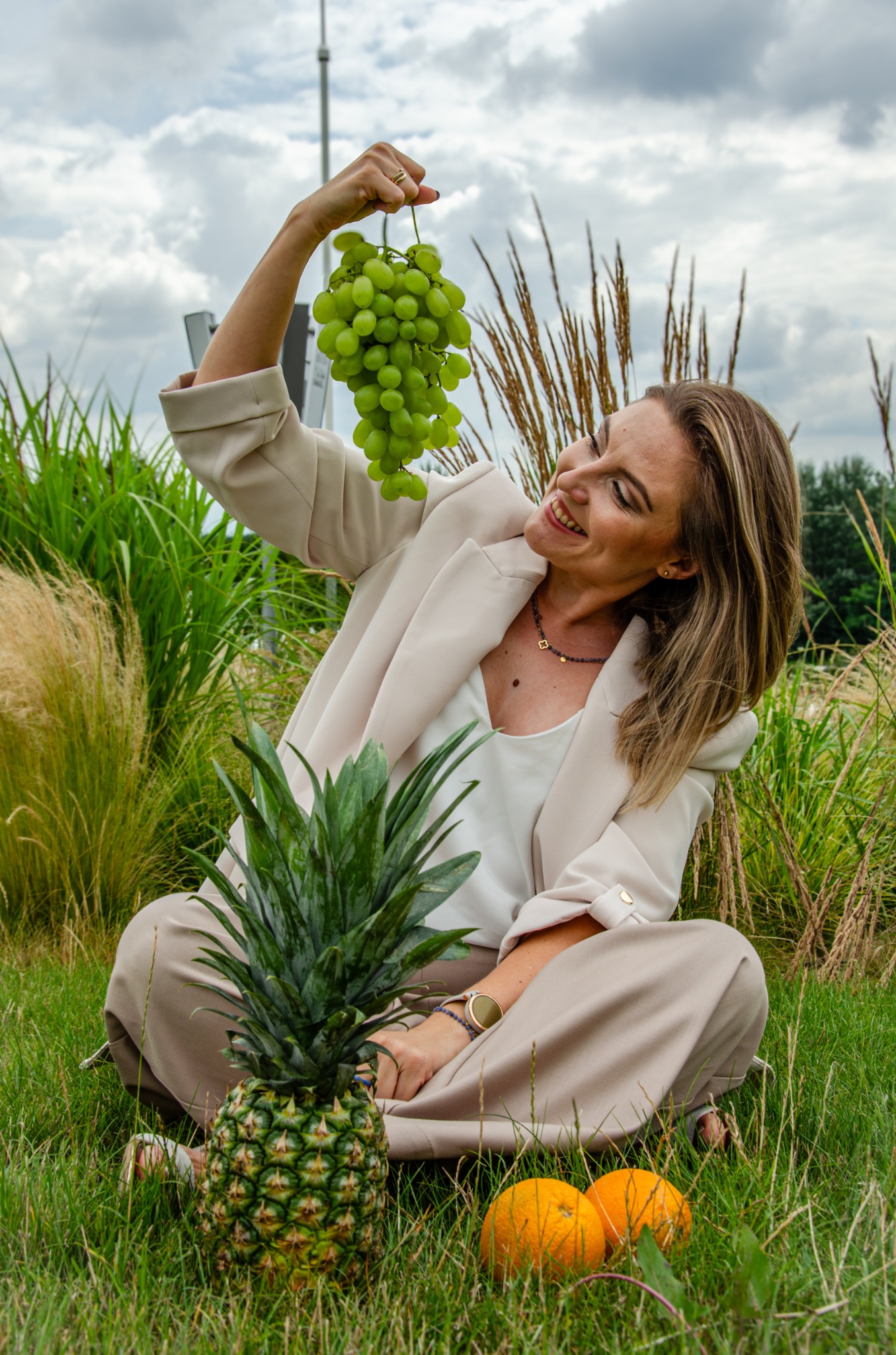 Uśmiechnięta kobieta w beżowym garniturze siedzi na trawie z ananasem i pomarańczami, trzymając nad głową kiść zielonych winogron