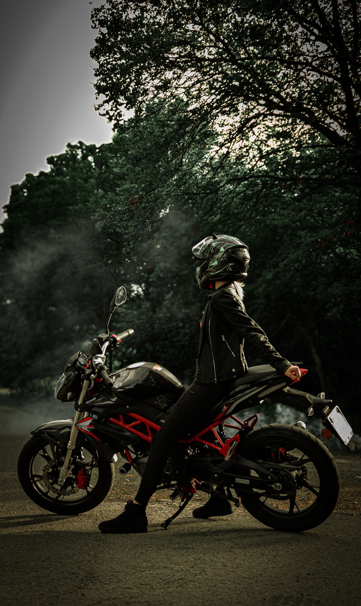 Osoba w czarnej kurtce i kasku siedzi na czarnym motocyklu Benelli z czerwoną ramą na tle drzew w Krakowie.