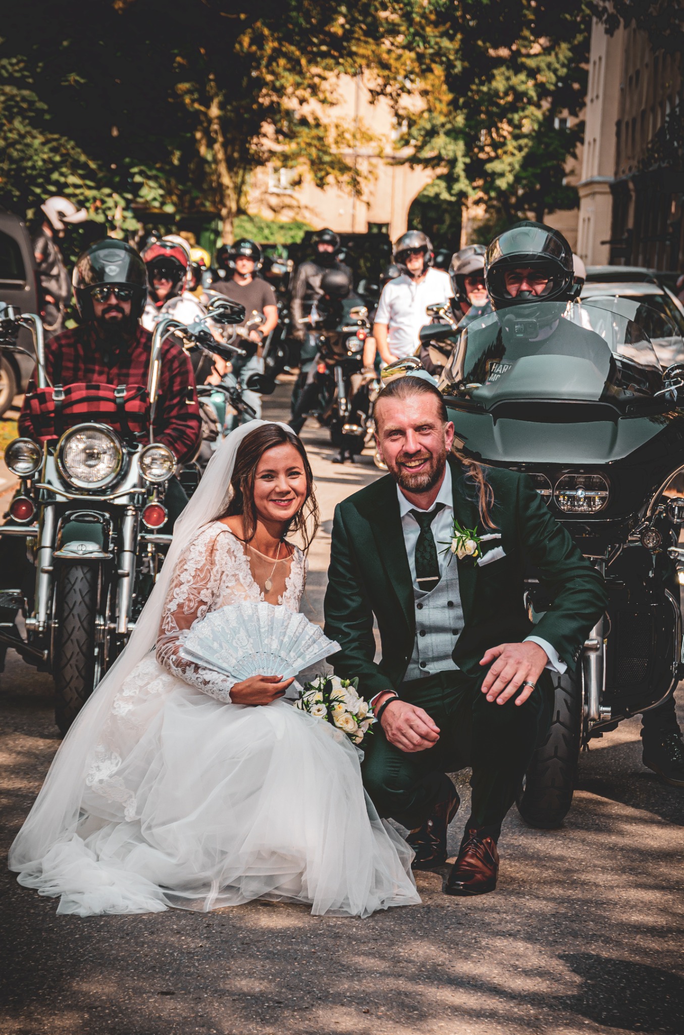 Uśmiechnięta para młoda w sukni ślubnej i zielonym garniturze, klęcząca na tle grupy motocyklistów i motocykli na ulicy miejskiej.