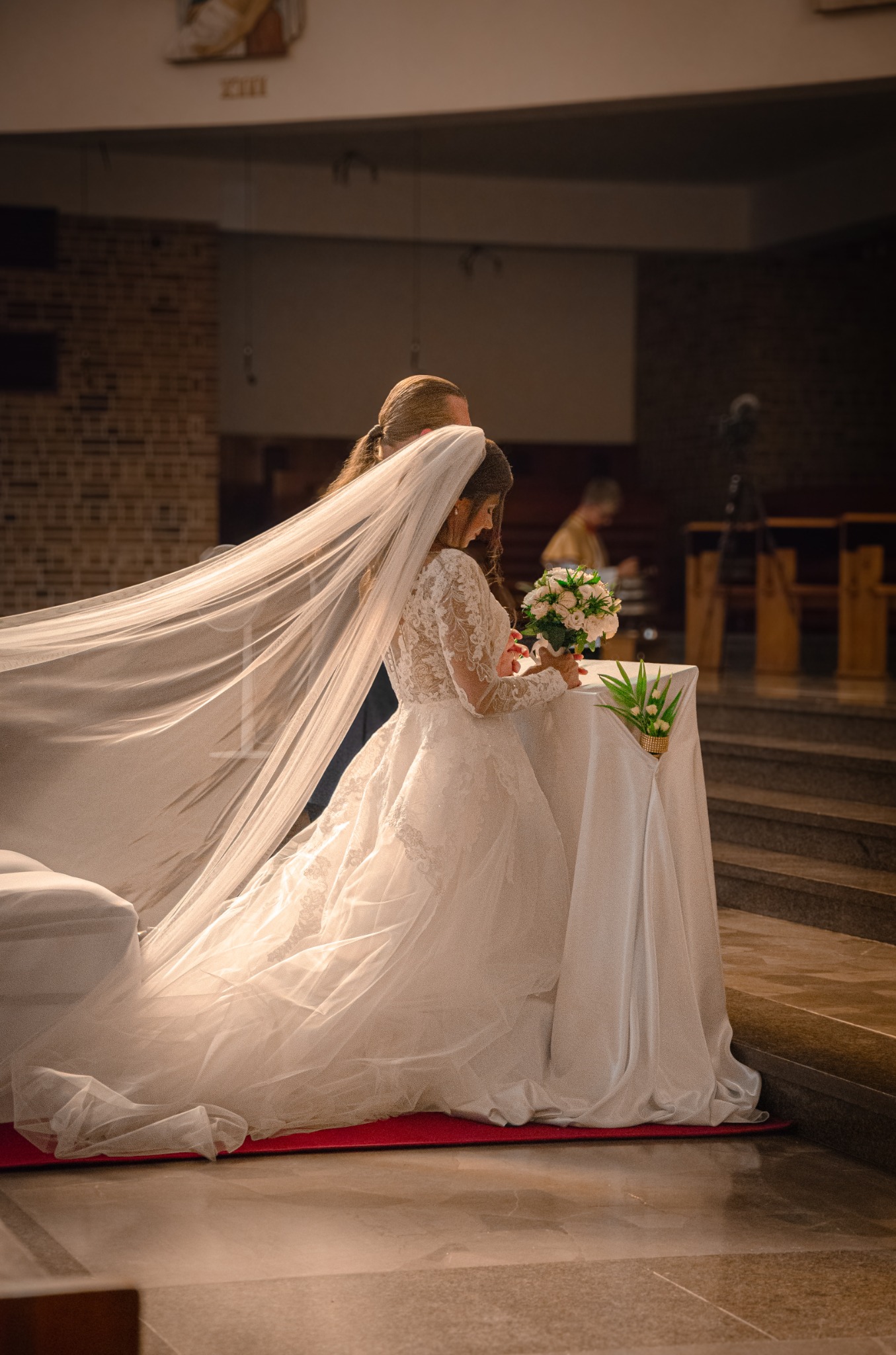 Panna młoda w koronkowej sukni ślubnej i długim welonie klęczy przy ołtarzu w kościele, trzymając bukiet kwiatów.