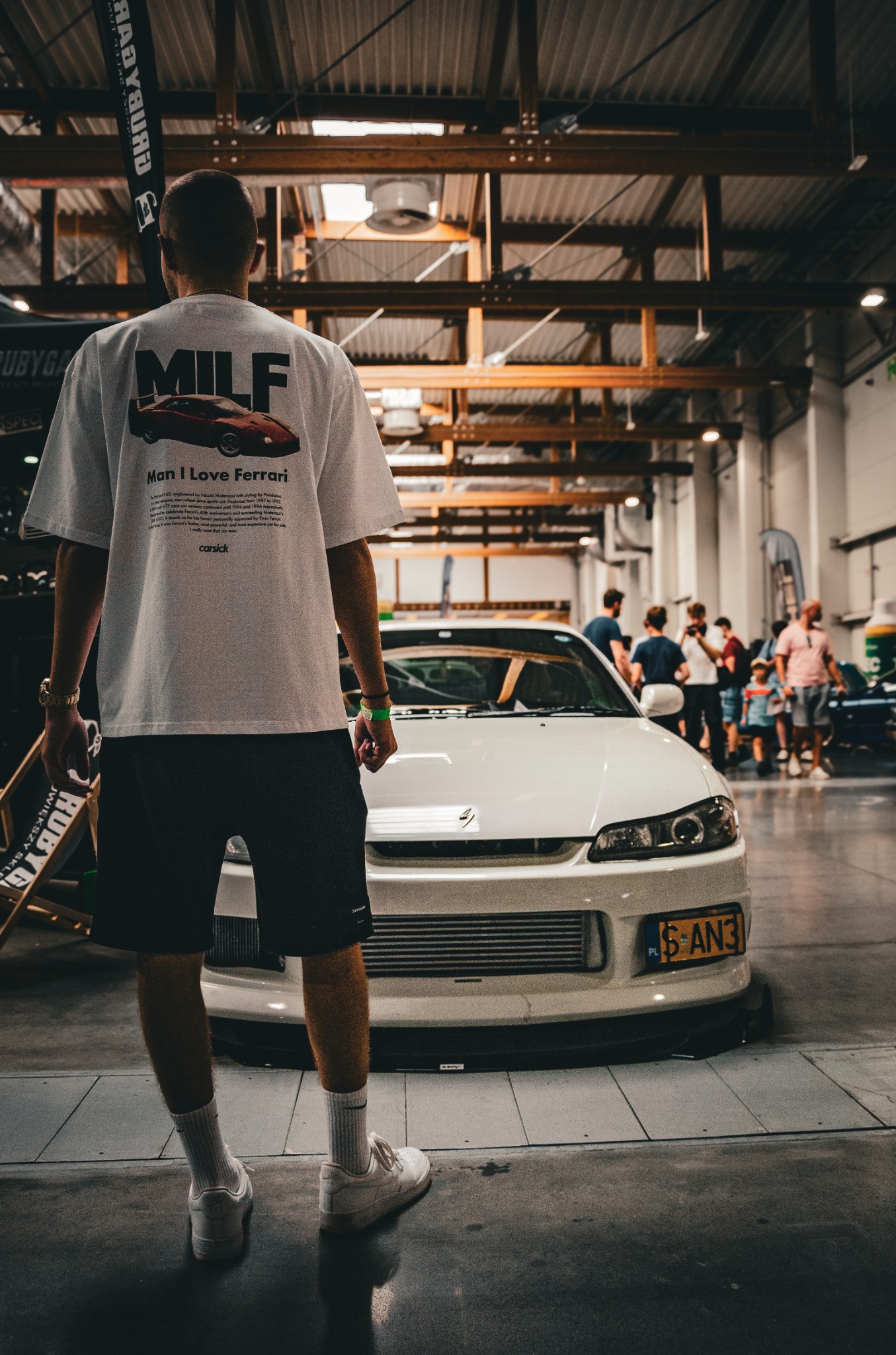 Mężczyzna w koszulce z nadrukiem Ferrari stojący przed białym sportowym samochodem Nissan Silvia S15 na hali wystawowej.