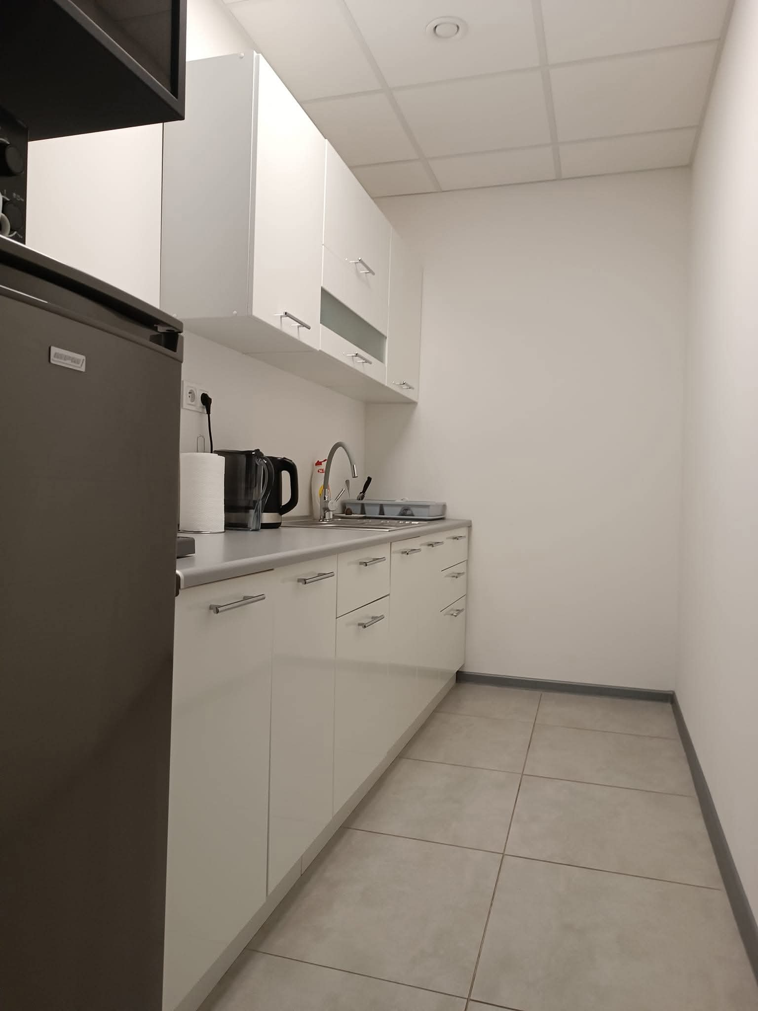 Wnętrze czystej, minimalistycznej kuchni biurowej z białymi szafkami, szarą podłogą i sprzętami AGD. Widoczny zlewozmywak, czajnik i dozownik płynu do naczyń.