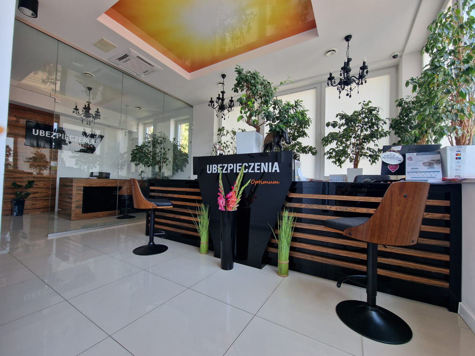 Nowoczesne biuro z recepcją z napisem 'Ubezpieczenia Optimum', drewnianymi krzesłami barowymi, roślinami doniczkowymi i podświetlanym sufitem.