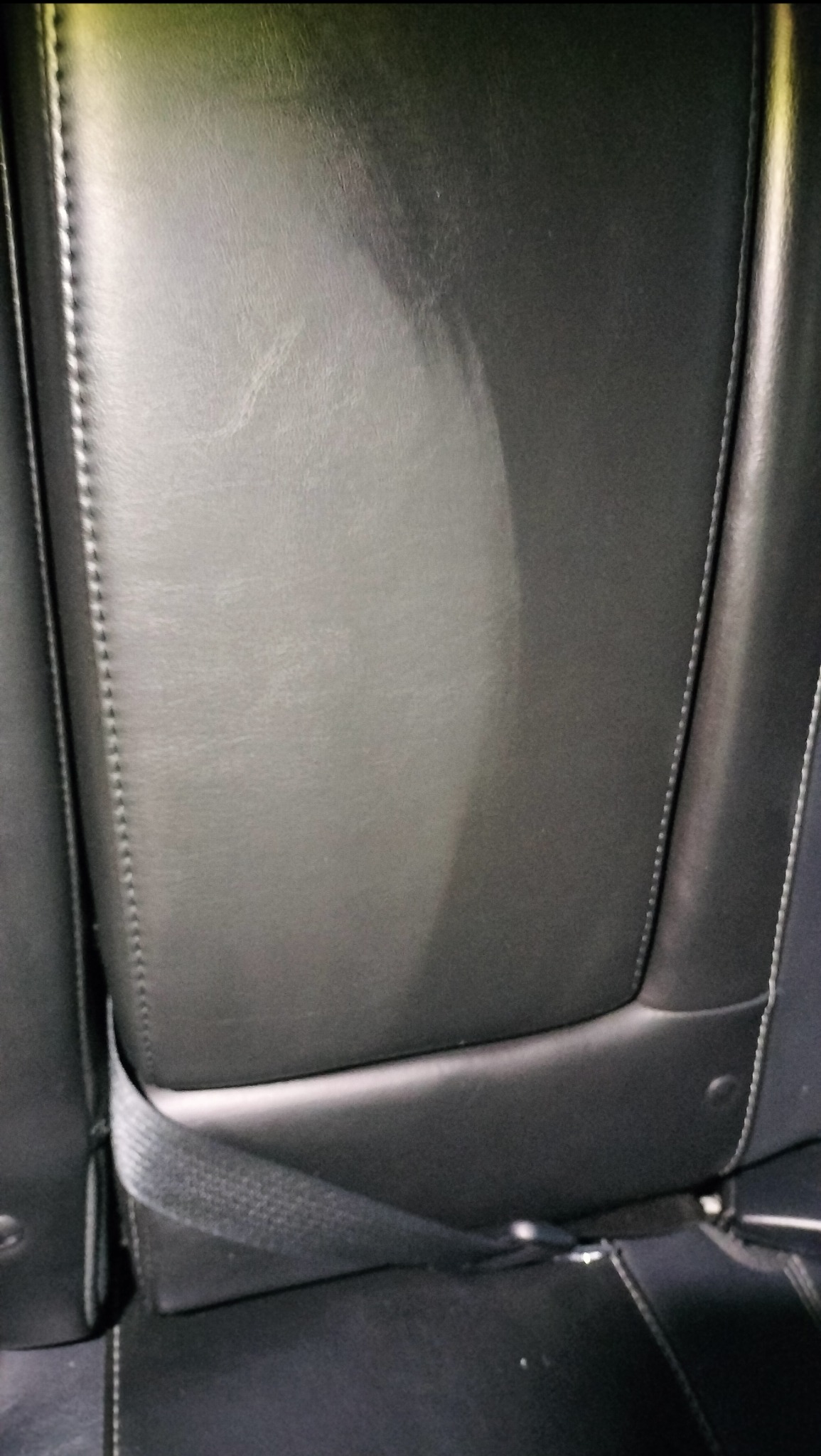 Fragment czarnego, skórzanego fotela samochodowego z widocznym pasem bezpieczeństwa i jaśniejszą plamą na oparciu, sugerującą świeżo wyczyszczoną powierzchnię.