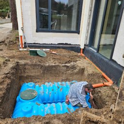 BB - Montaż niebieskiego zbiornika na szambo w wykopie obok budynku z oknami, widoczny pracownik w pasiastej koszuli, rury kanalizacyjne i ramię koparki Bobcat.