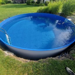 BB - Okrągły basen częściowo wkopany w ziemię, wypełniony wodą, drabinka przymocowana, widoczne buty osoby w tle, trawa i budynek mieszkalny.