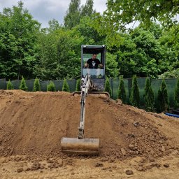 BB - Operator w minikoparce pracuje na kopcu ziemi, w tle widoczne tuje i zielone drzewa.