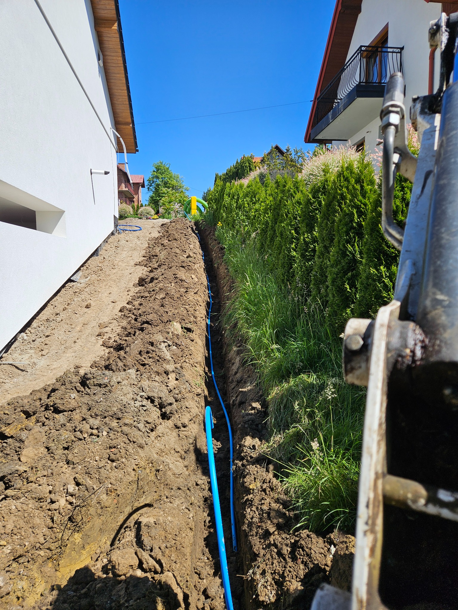Wykopany wąski rów z niebieskimi rurami na dnie, wykonany wzdłuż granicy działki z widocznym fragmentem koparki i domami w tle.