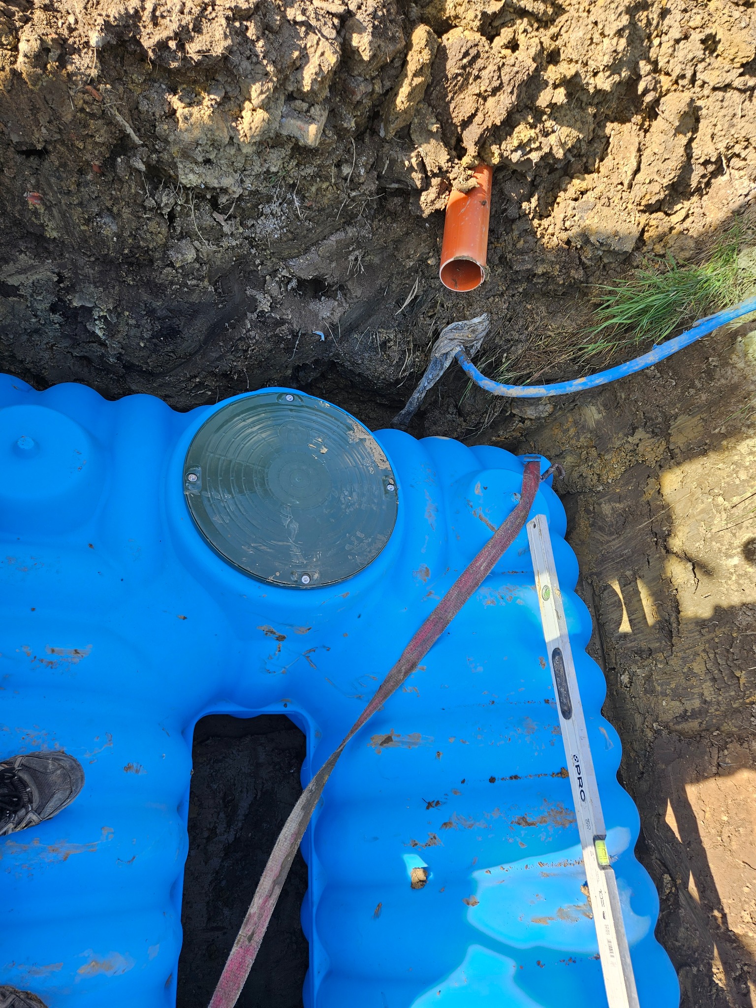 Niebieski, plastikowy szambo zakopany w ziemi, widoczny wykop, rura kanalizacyjna, poziomica i fragment buta na krawędzi szamba.