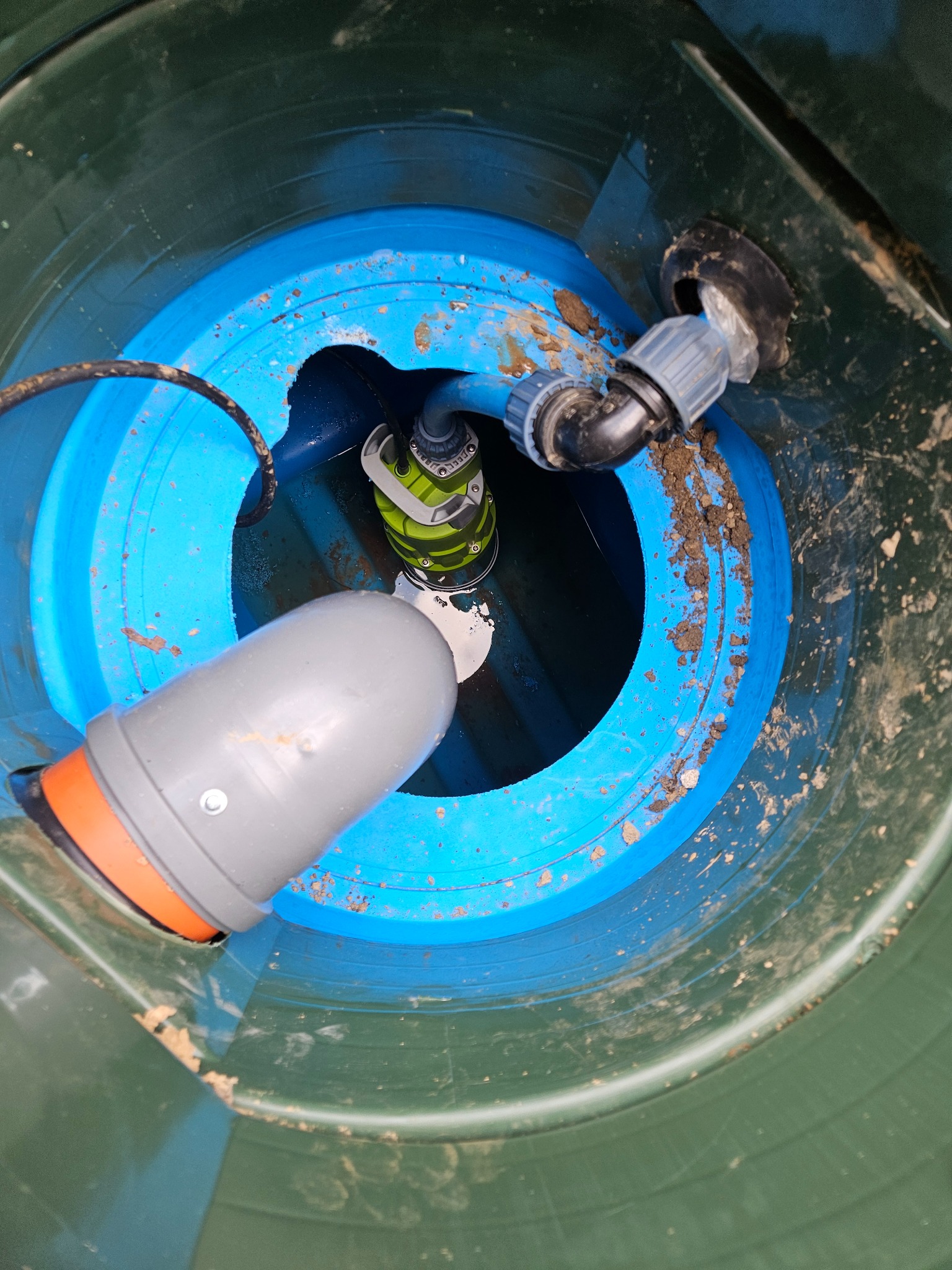 Wnętrze zielonego szamba z niebieskim pierścieniem, widoczna pompa zanurzeniowa z zielonymi akcentami, szare rury odpływowe z pomarańczowym uszczelnieniem i brudne ściany zbiornika.