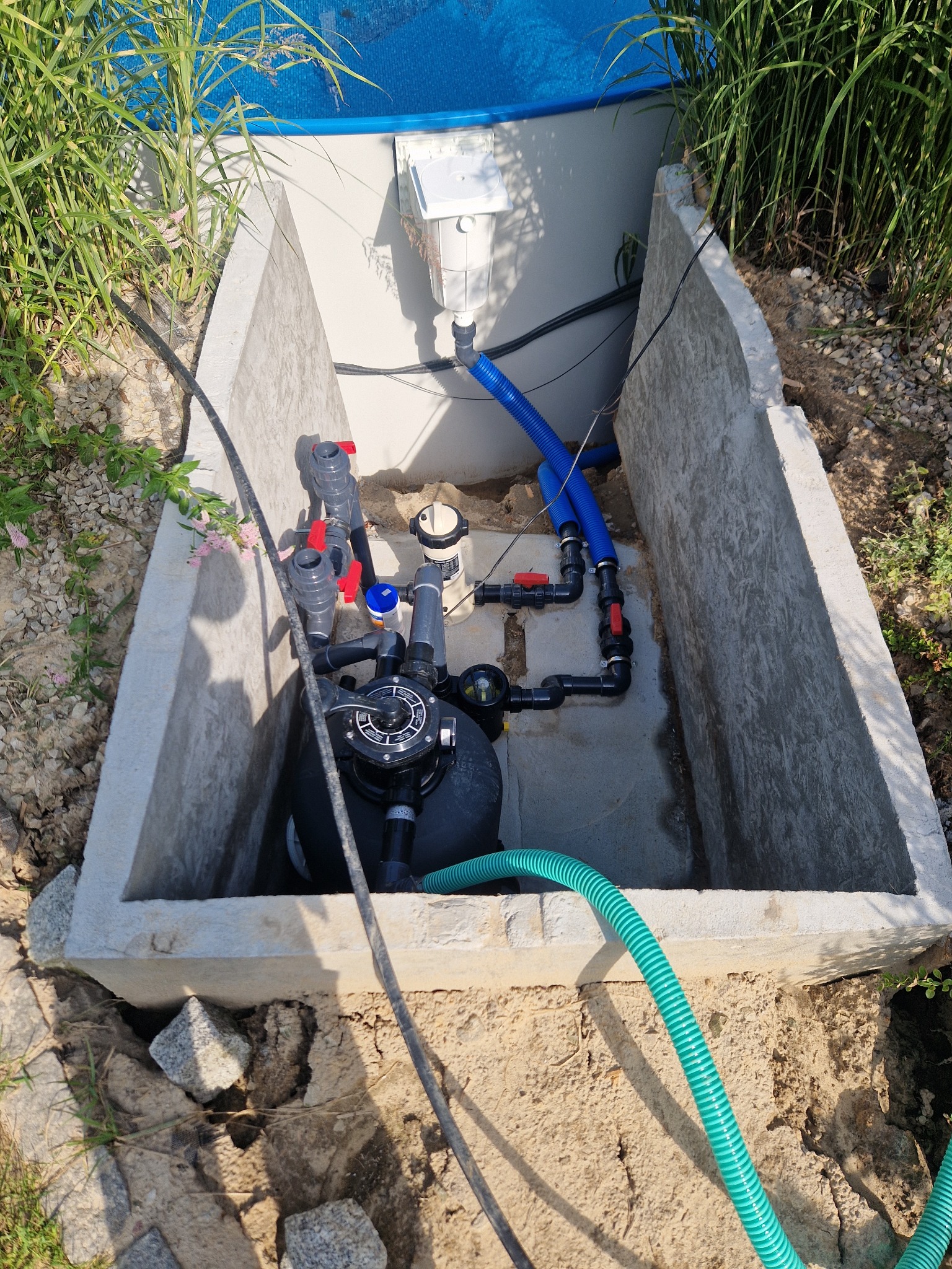 Betonowa studzienka z filtrem basenowym, rurami PCV i zielonym wężem, otoczona roślinnością i kamieniami.