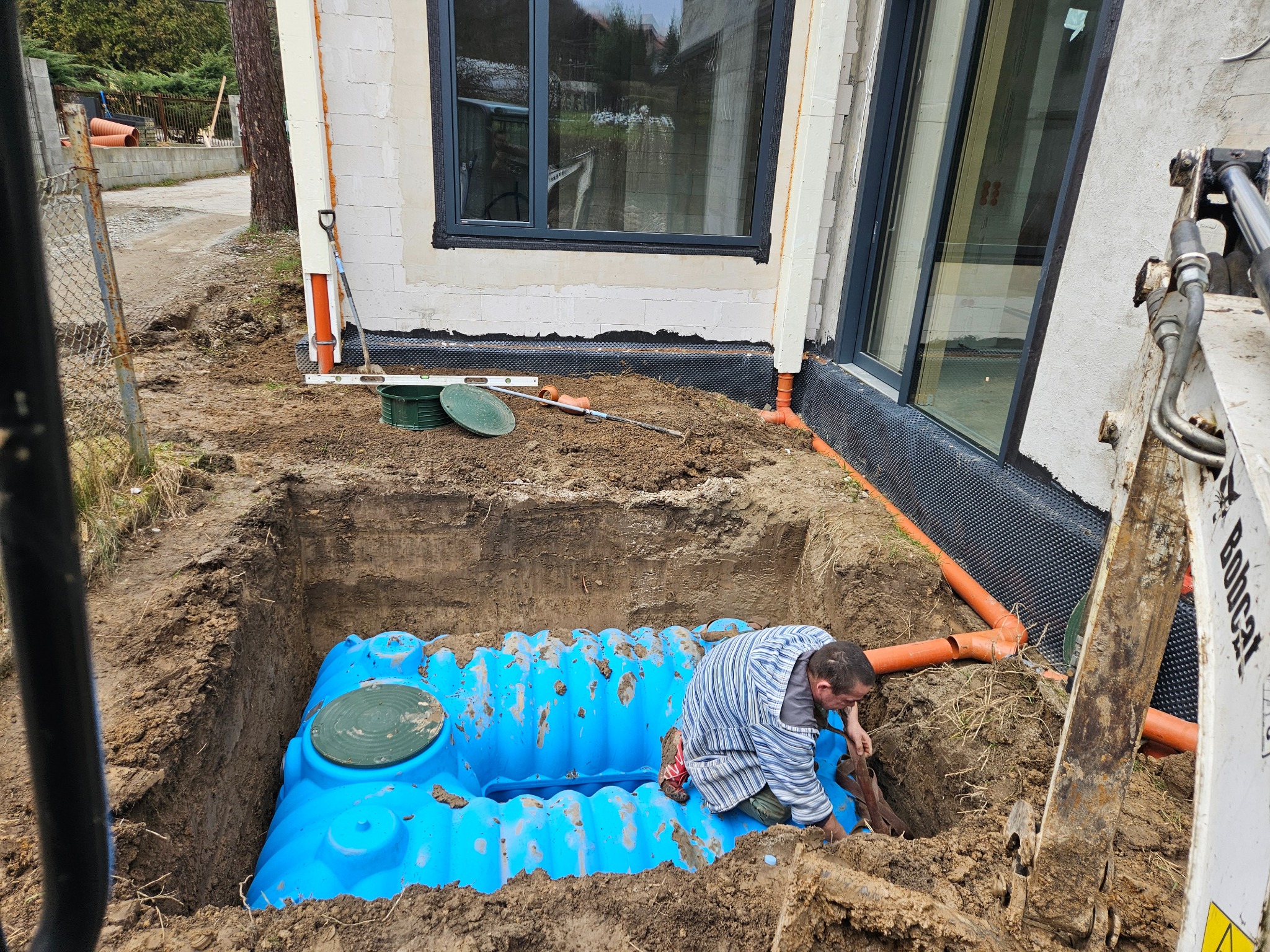 Montaż niebieskiego zbiornika na szambo w wykopie obok budynku z oknami, widoczny pracownik w pasiastej koszuli, rury kanalizacyjne i ramię koparki Bobcat.