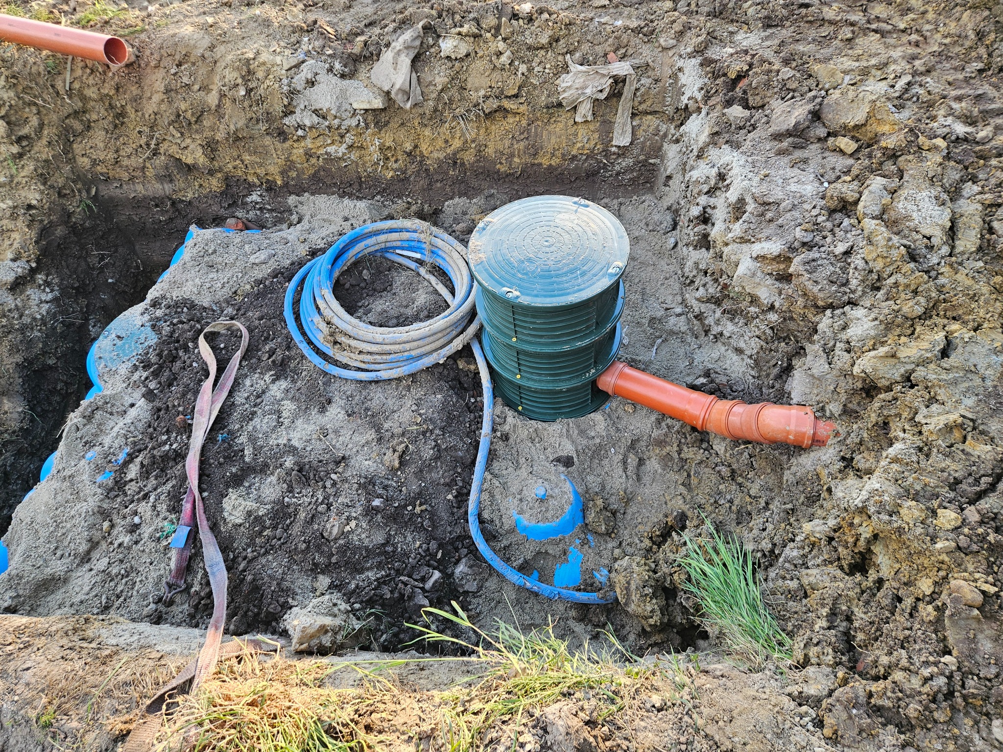 Zielony zbiornik na szambo umieszczony w wykopie, widoczne rury kanalizacyjne i niebieski kabel, ziemia i trawa wokół wykopu.