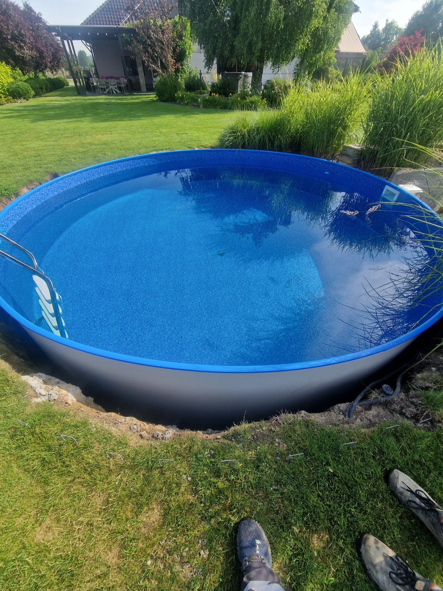 Okrągły basen częściowo wkopany w ziemię, wypełniony wodą, drabinka przymocowana, widoczne buty osoby w tle, trawa i budynek mieszkalny.