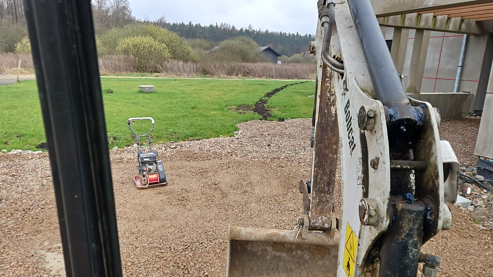 Widok z kabiny koparki na teren z utwardzoną żwirową nawierzchnią, ubijarkę wibracyjną i wykopaną bruzdę w trawie prowadzącą do budynku w tle.