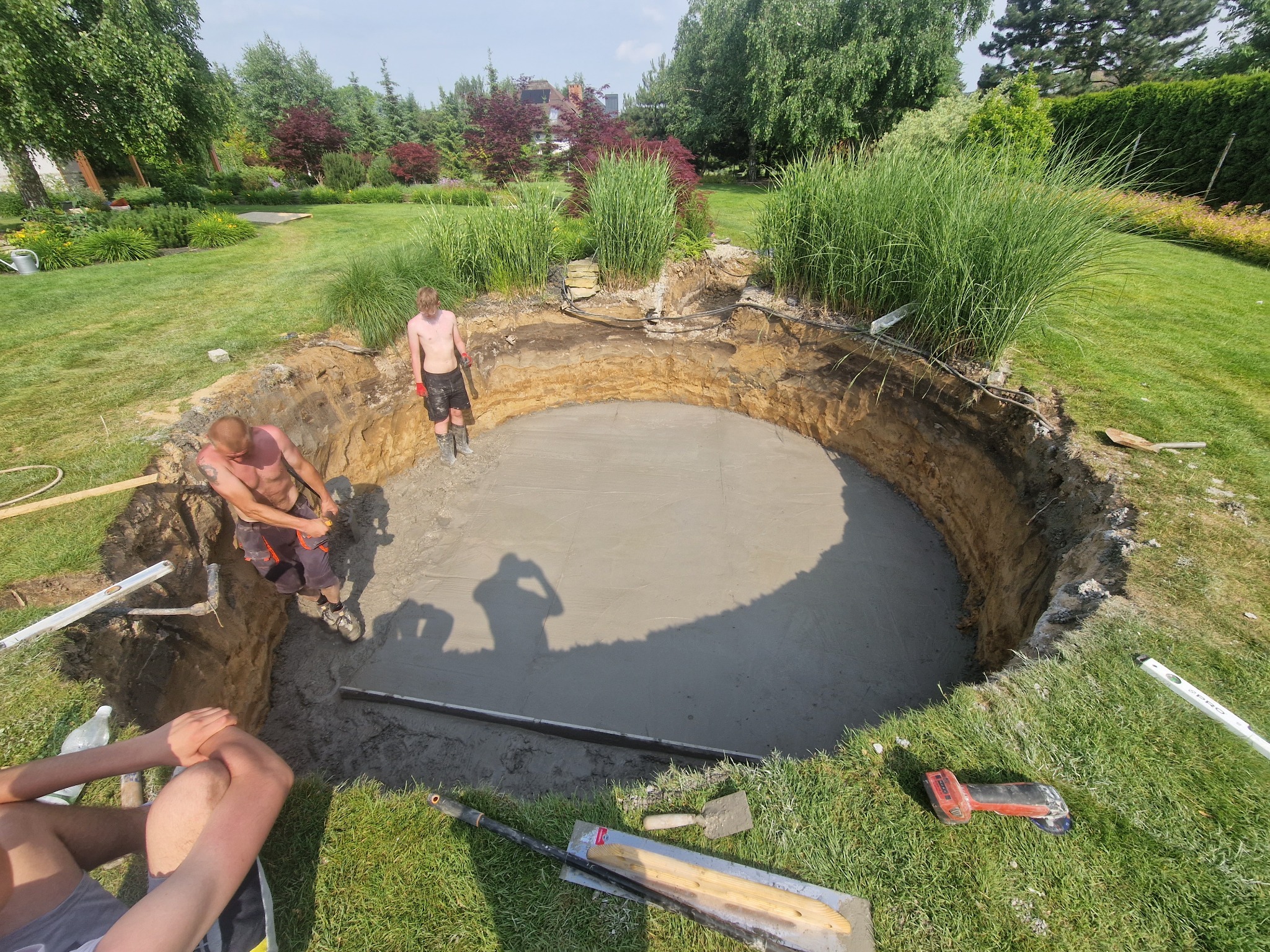 Okrągły wykop w ziemi z wylanym betonem na dnie, dwóch mężczyzn w środku i na krawędzi, narzędzia budowlane na trawie wokół, trawa, krzewy i drzewa w tle.