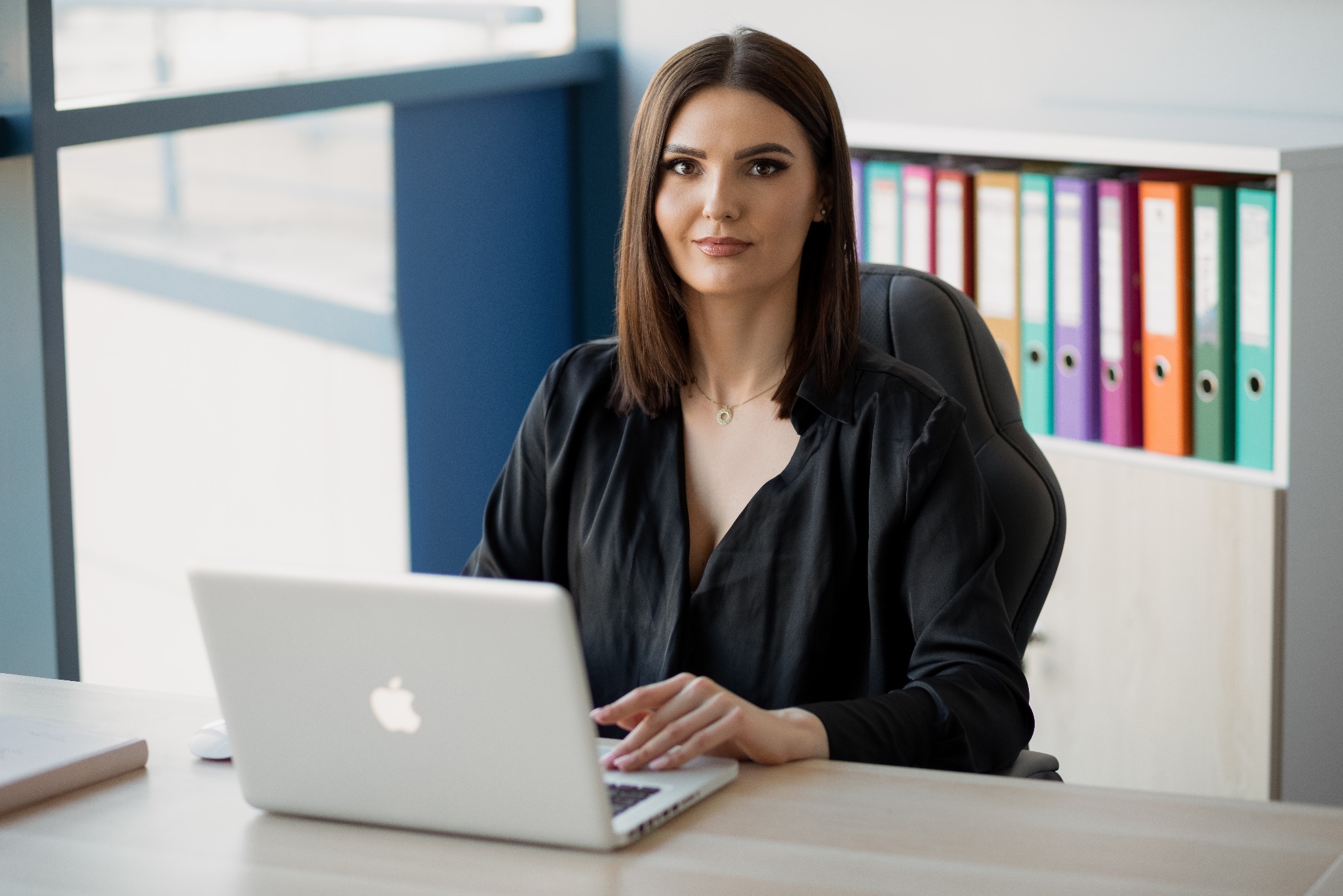 Kobieta w czarnej koszuli siedzi za biurkiem i pracuje na laptopie, w tle regał z kolorowymi segregatorami.