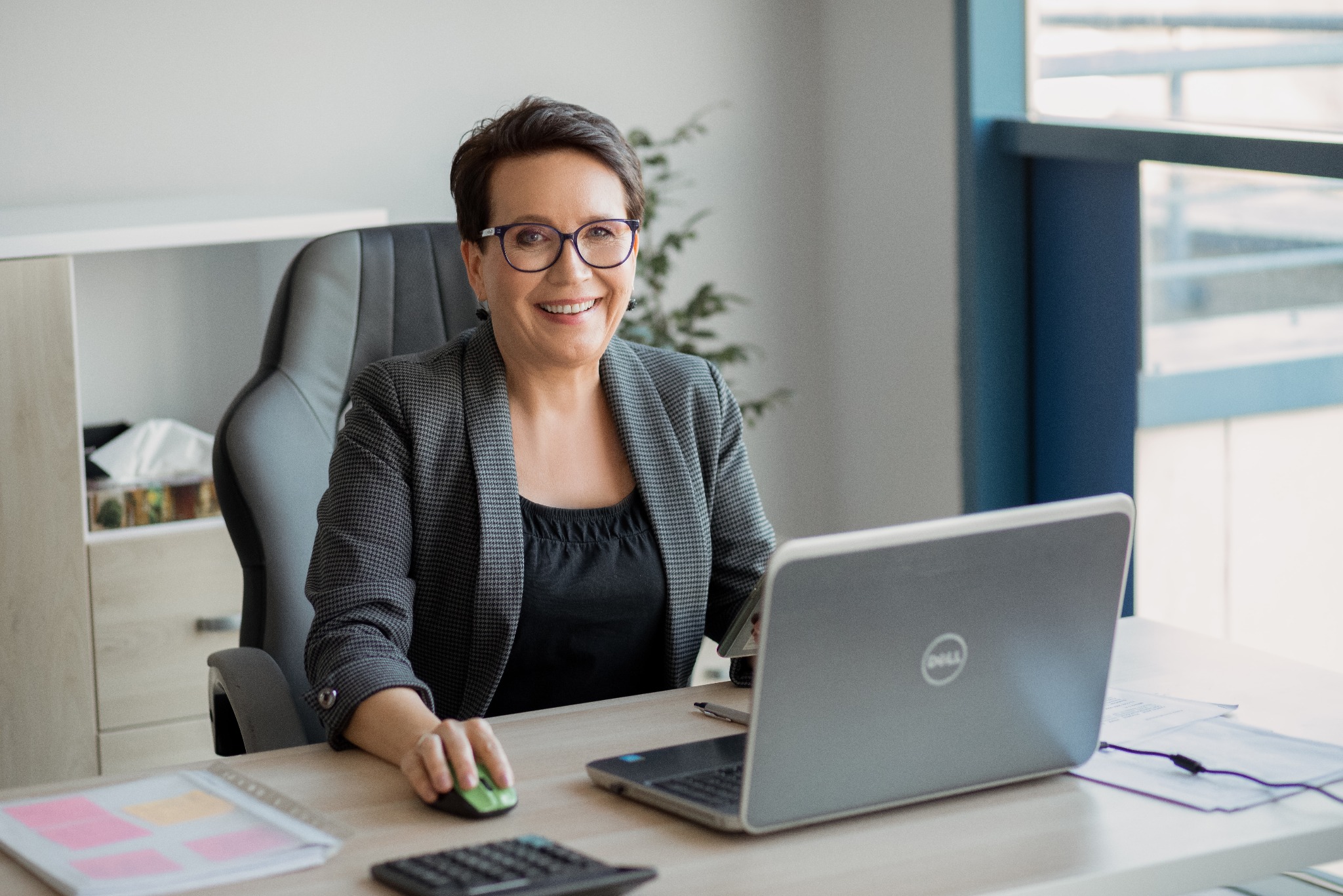 Uśmiechnięta kobieta w okularach i szarej marynarce siedzi przy biurku z laptopem Dell, kalkulatorem i notatkami.