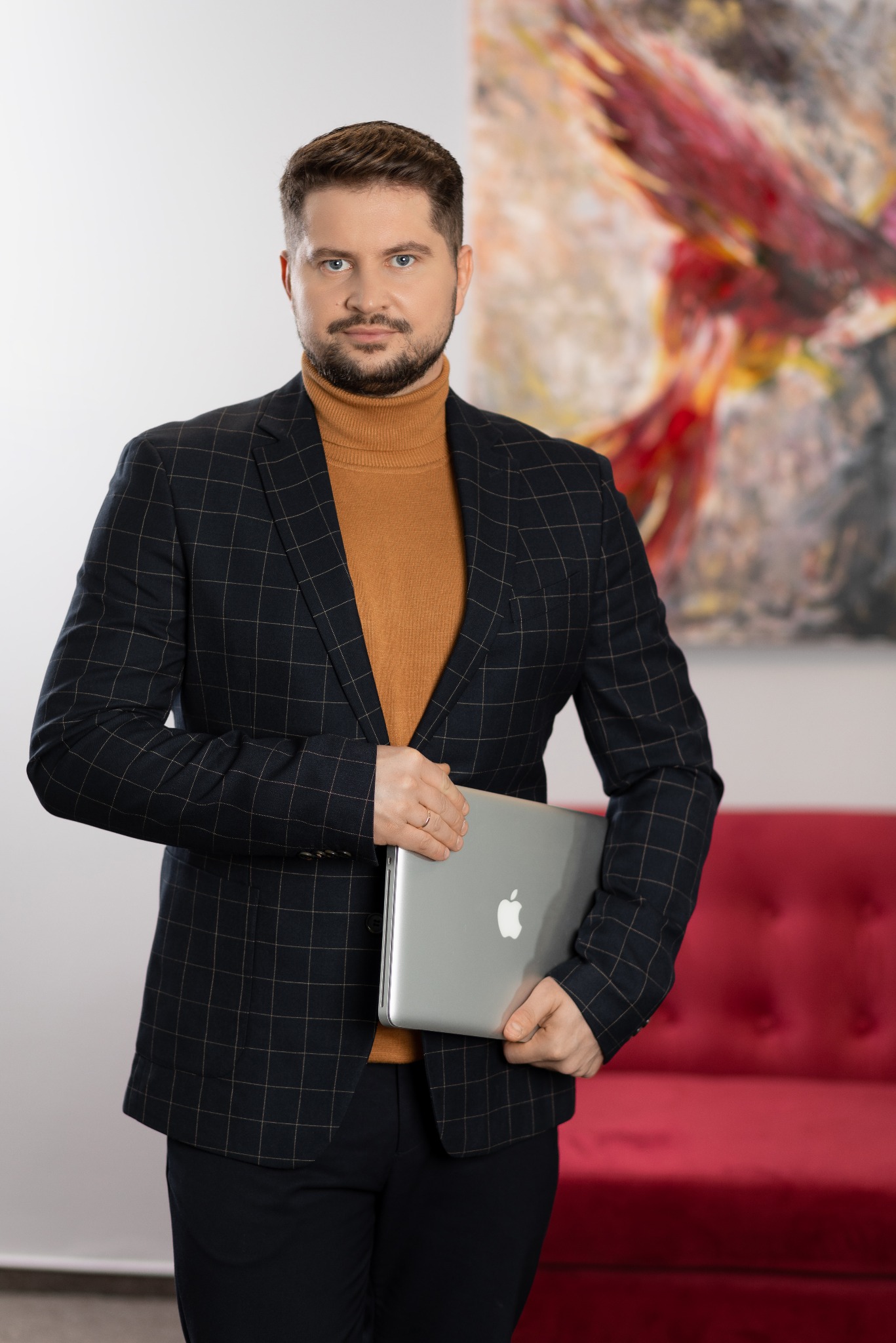 Mężczyzna w marynarce w kratę i golfie trzyma laptopa na tle abstrakcyjnego obrazu i czerwonej sofy.