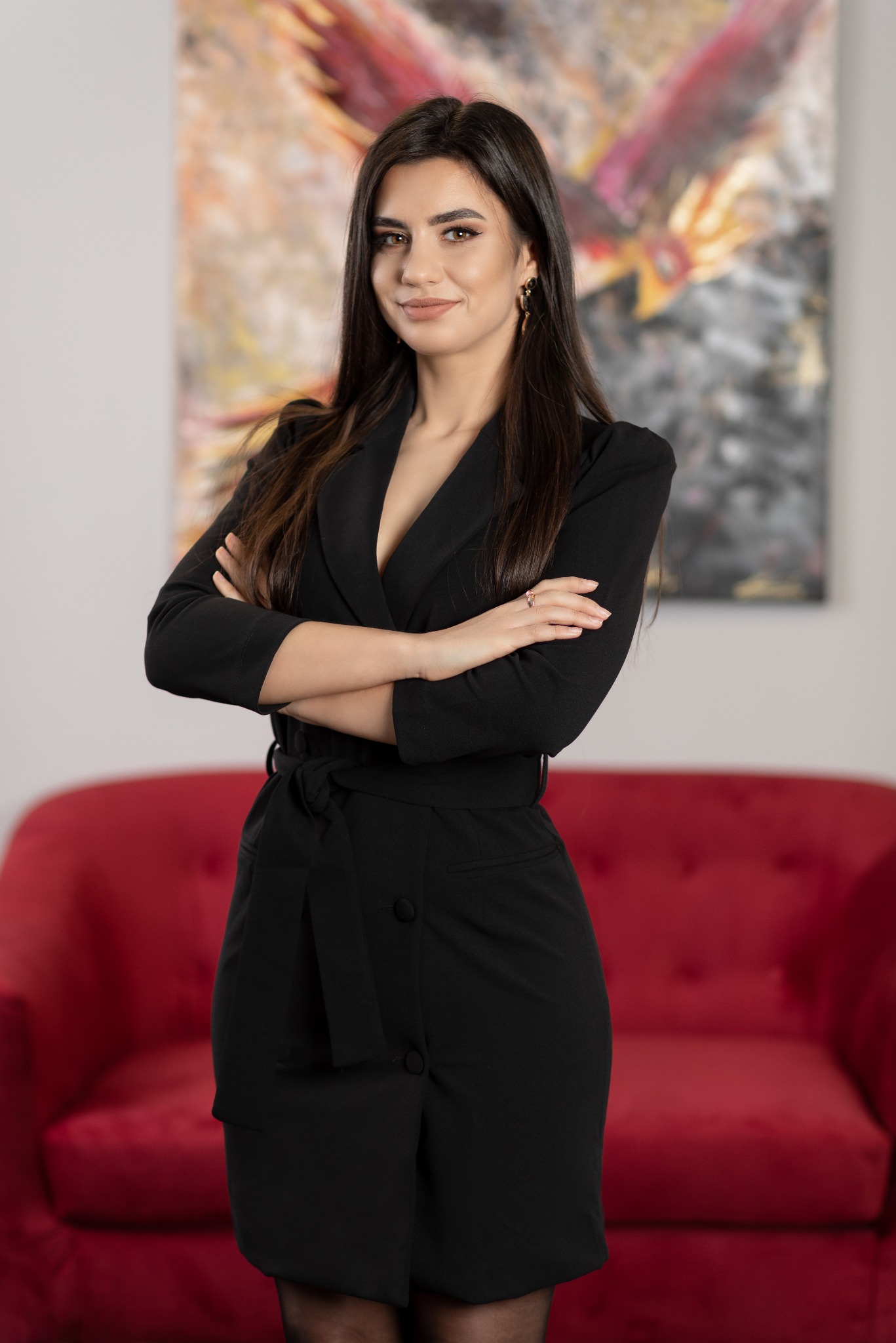 Portret kobiety w czarnym żakiecie z rękami skrzyżowanymi na tle czerwonej sofy i abstrakcyjnego obrazu.
