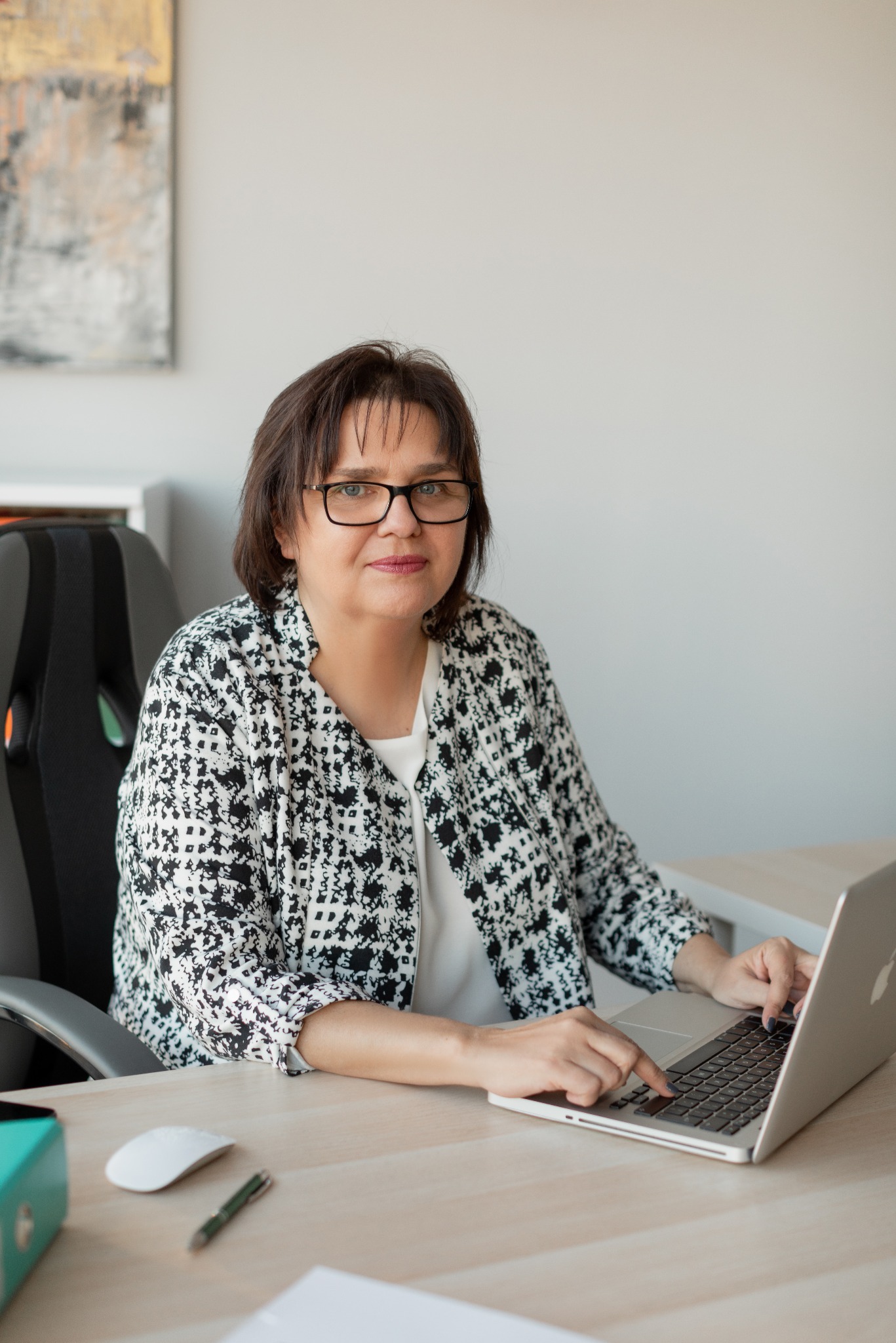 Kobieta w okularach i wzorzystej marynarce pracuje na laptopie przy biurku w jasnym gabinecie; w tle abstrakcyjny obraz.