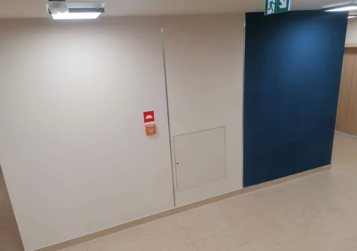 Korytarz z pomalowaną na granatowo ścianą, białą ścianą z klapą rewizyjną i czujnikiem dymu, oraz płytkami podłogowymi w odcieniach beżu.