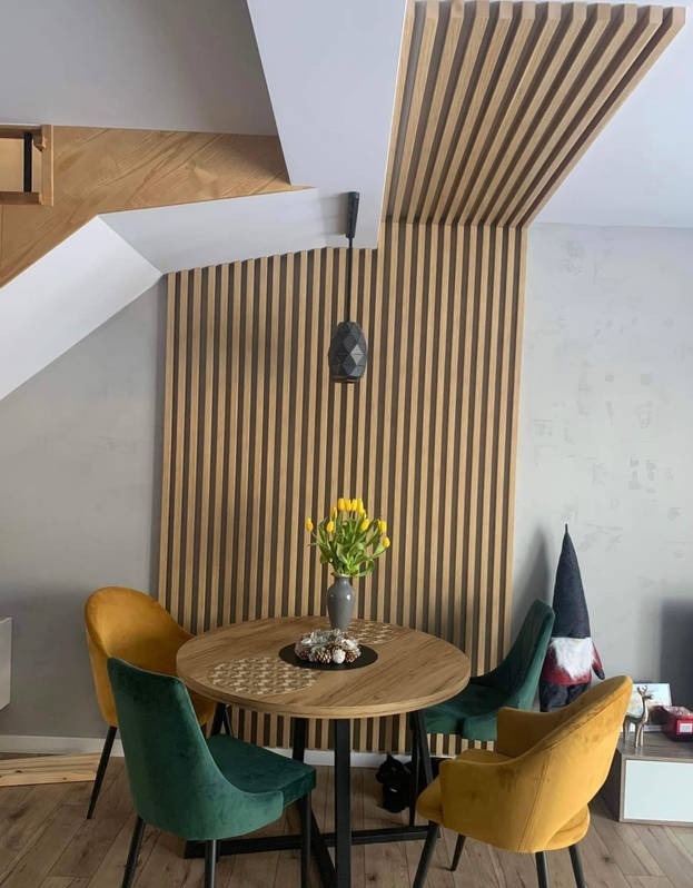 Jadalnia z drewnianą lamelową ścianą i sufitem, okrągły stół z tulipanami w wazonie, zielone i musztardowe krzesła, dekoracja świąteczna w tle.