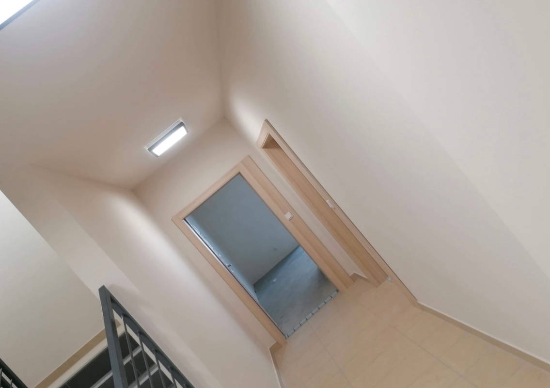 Klatka schodowa w jasnych barwach, z widocznym fragmentem balustrady, świeżo pomalowane ściany oraz futryna drzwiowa z zamontowanymi drzwiami, oświetlenie sufitowe w postaci panelu LED.