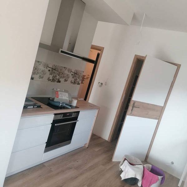 Montaż mebli kuchennych: białe szafki z piekarnikiem, płyta indukcyjna, okap oraz częściowo zamontowane drzwi w ościeżnicy, widoczne akcesoria montażowe na blacie.