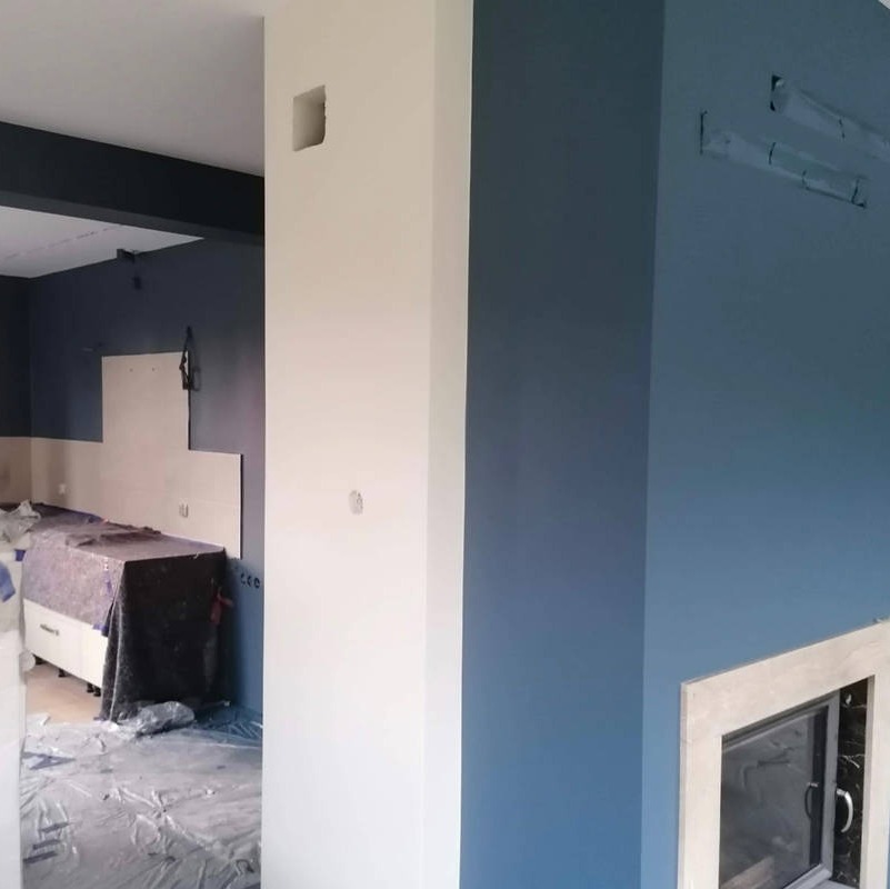 Wnętrze w trakcie malowania: biały filar kontrastuje z granatową ścianą, widoczne zabezpieczenia podłogi i fragment kominka z jasną obudową.