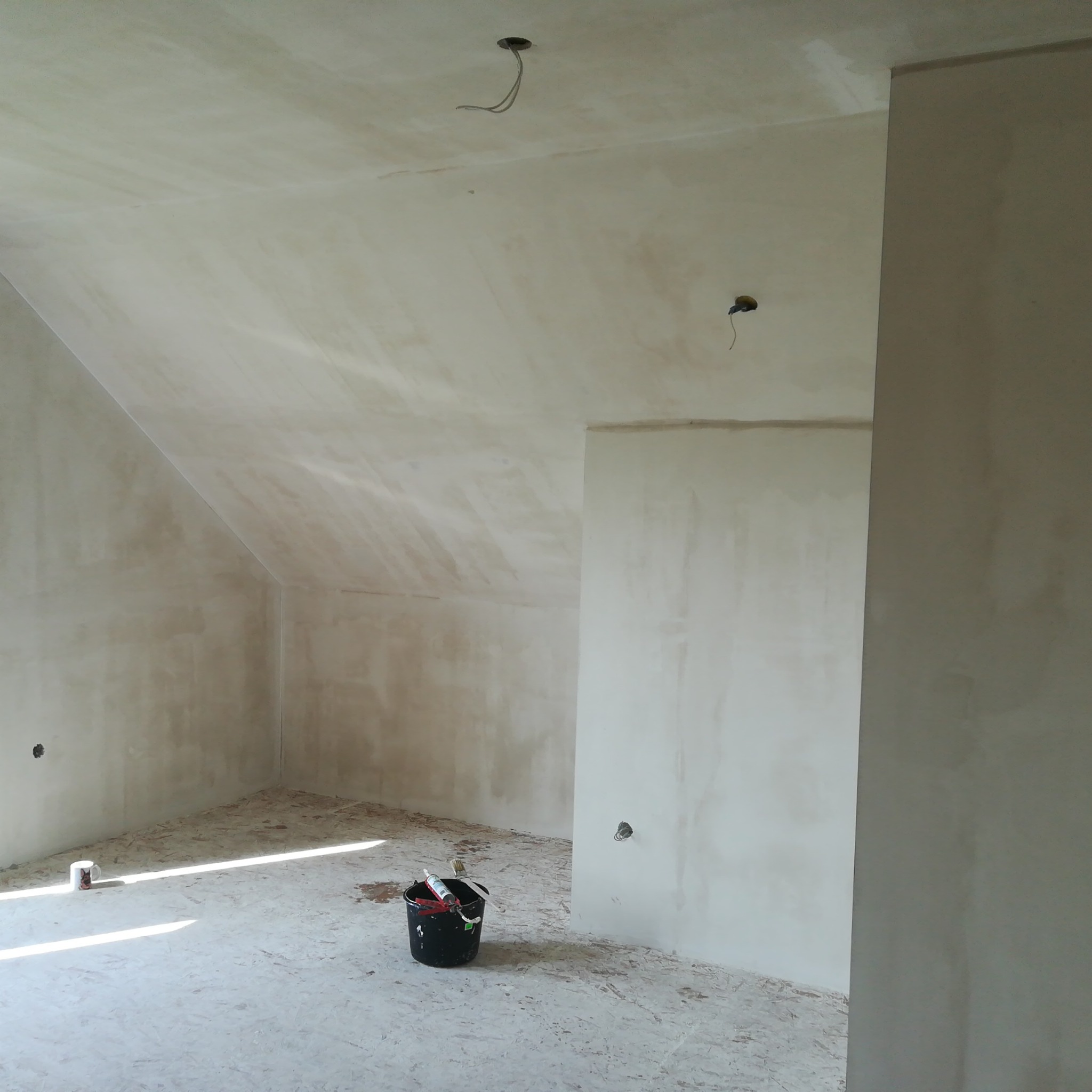 Pokój na poddaszu w trakcie prac wykończeniowych, ściany pokryte świeżo nałożoną gładzią, widoczne puszki elektryczne i wiadro z narzędziami na podłodze.