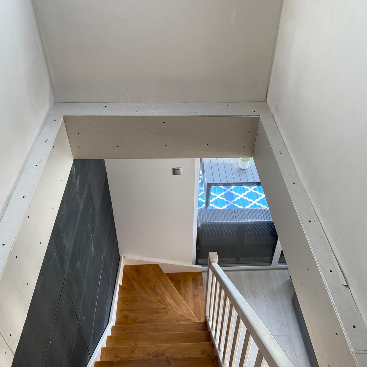 Klatka schodowa w trakcie prac wykończeniowych: widoczne płyty kartonowo-gipsowe na ścianach i suficie, drewniane schody, czarna ściana, widok na taras z niebieskim wzorem.