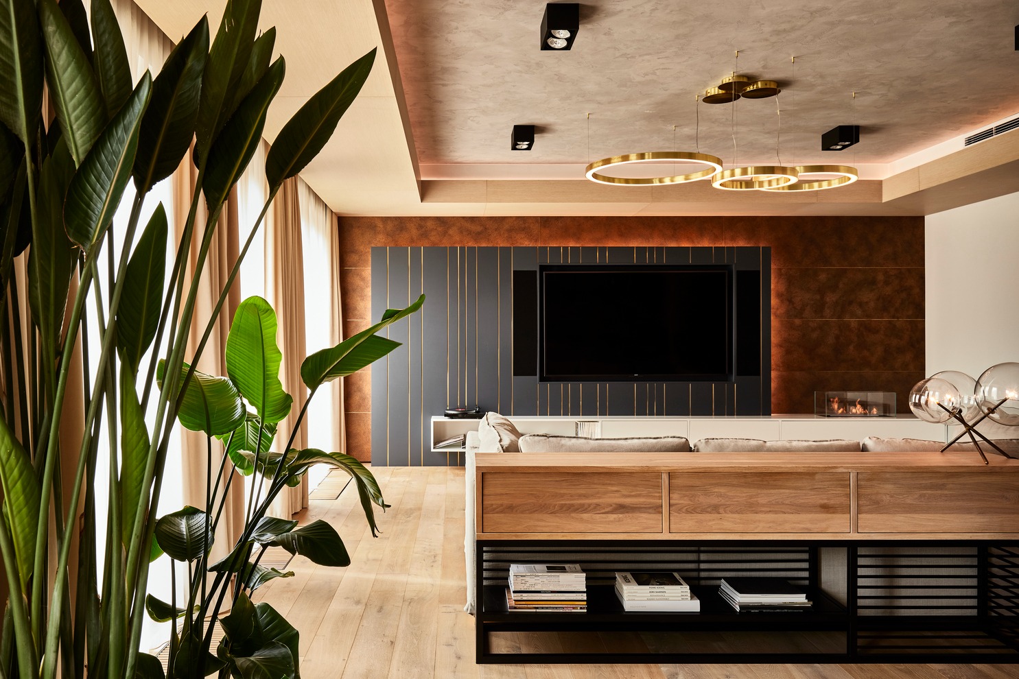 Salon z drewnianą podłogą, ścianą akcentową z telewizorem, rośliną doniczkową i okrągłymi lampami wiszącymi, oświetlony ciepłym światłem.