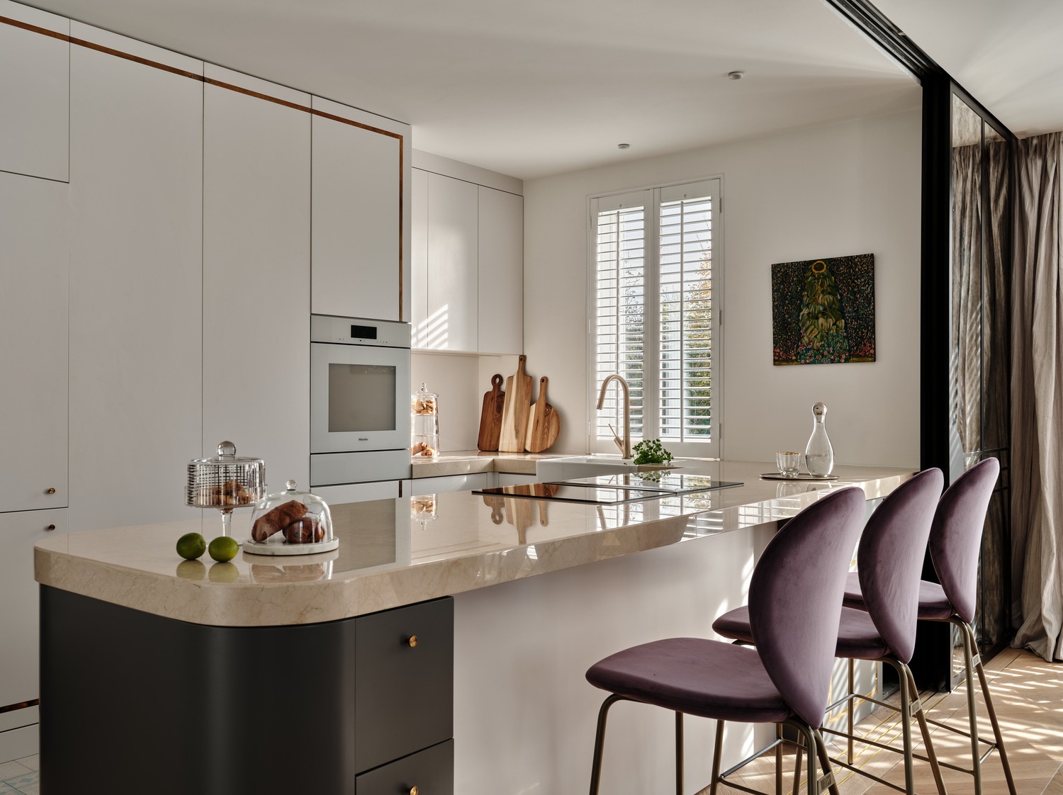 Elegancka kuchnia z wyspą o marmurowym blacie, nowoczesnymi białymi szafkami z miedzianymi akcentami, szarym dołem wyspy, piekarnikiem Miele, trzema fioletowymi krzesłami barowymi i dziełem sztuki...