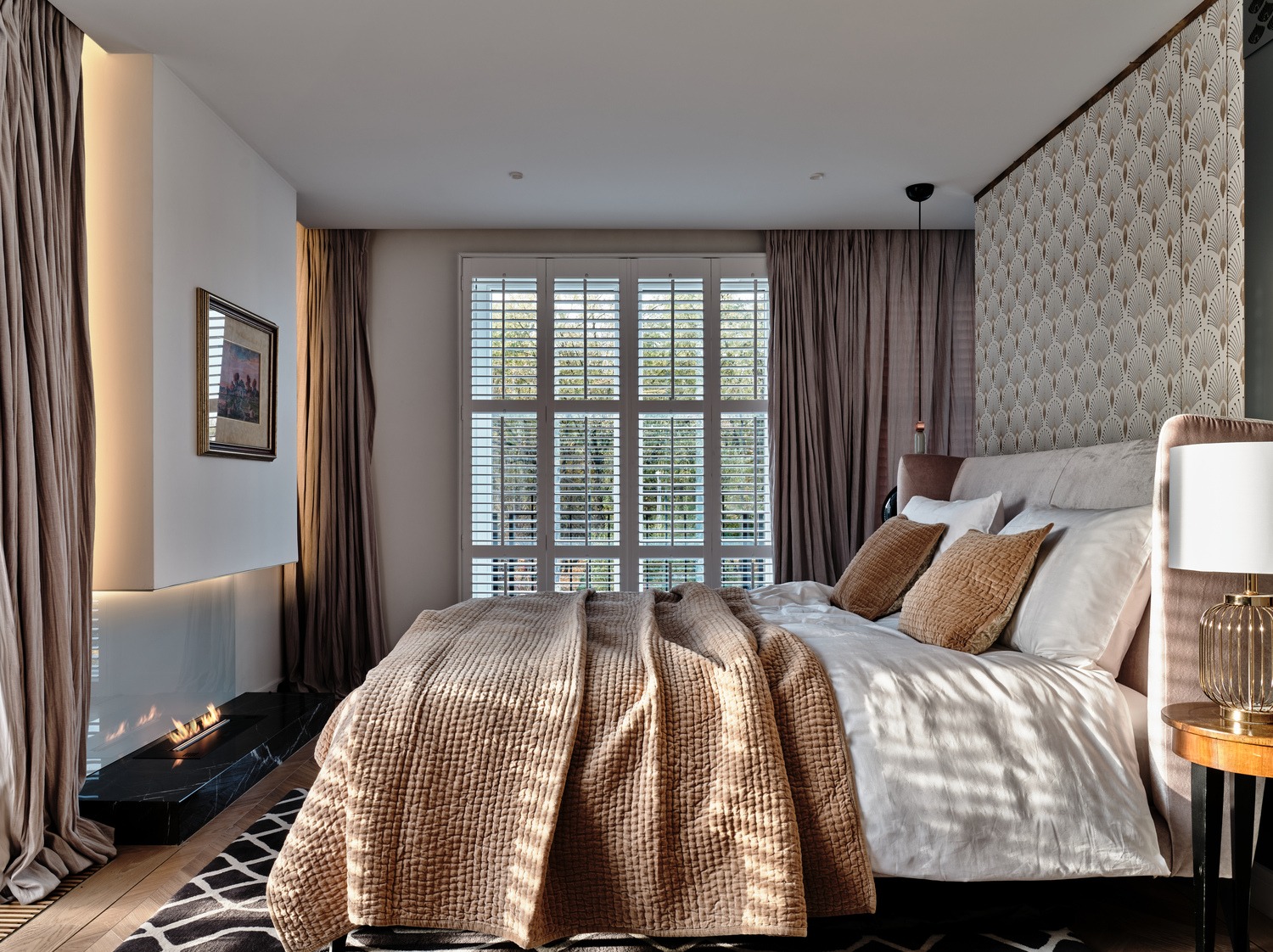 Elegancka sypialnia z kominkiem bio, zasłonami w odcieniach beżu i białymi żaluzjami okiennymi, uzupełniona wzorzystą tapetą za wezgłowiem łóżka i geometrycznym dywanem.