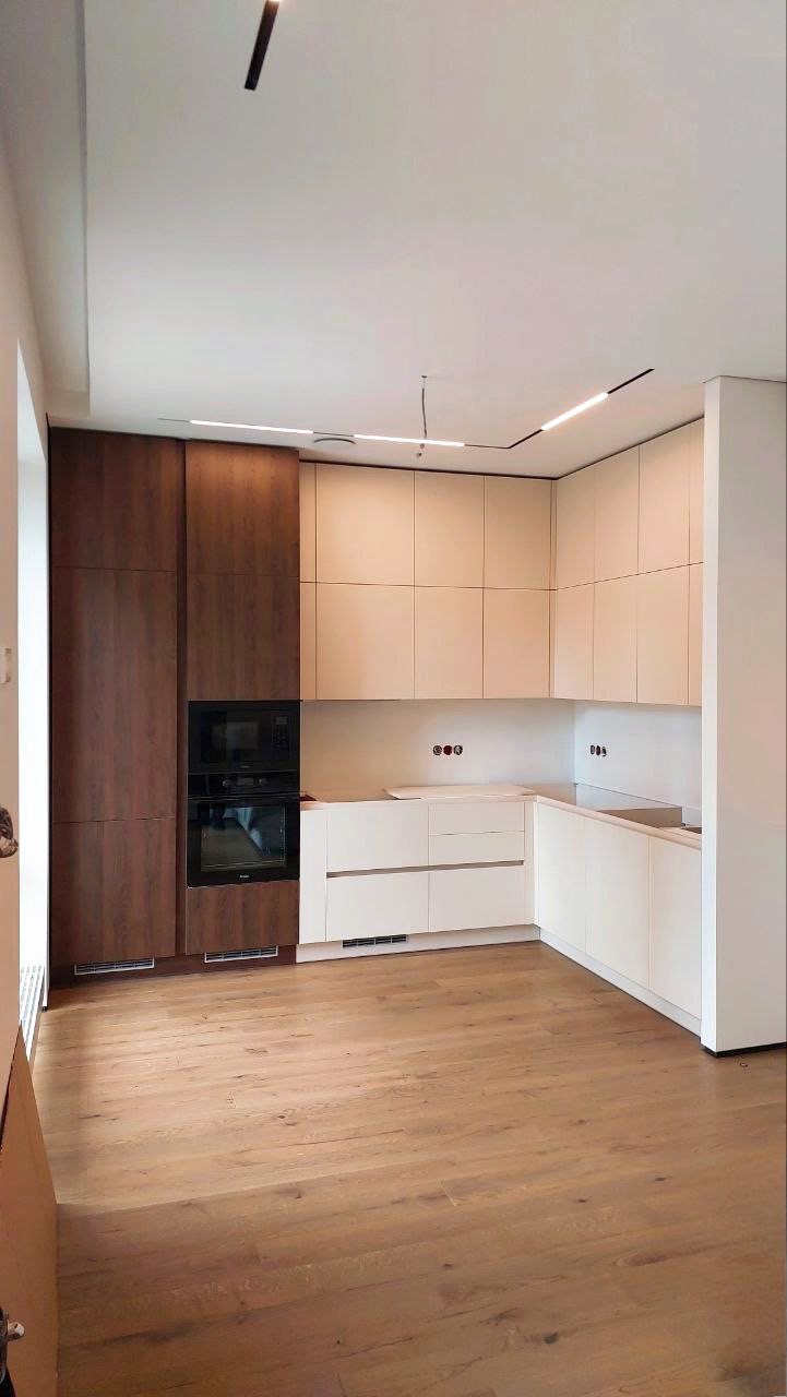 Nowoczesna kuchnia w Krakowie z białymi i drewnianymi szafkami, zintegrowanym piekarnikiem i mikrofalówką, oraz drewnianą podłogą. Oświetlenie sufitowe liniowe.