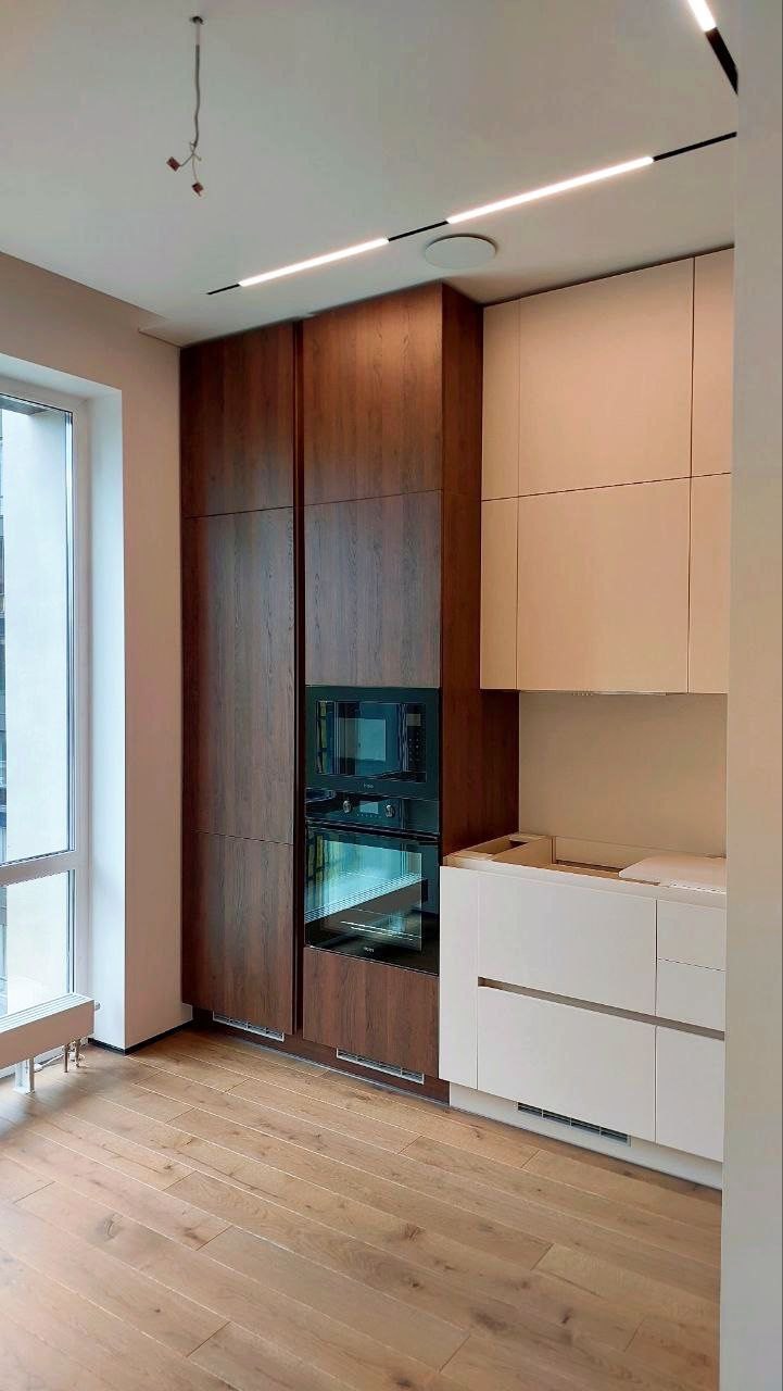 Nowoczesna kuchnia z zabudową meblową w odcieniach brązu i bieli, z wbudowanym piekarnikiem i mikrofalówką, oświetleniem ledowym i drewnianą podłogą, widoczne niedokończone podłączenie oświetlenia...