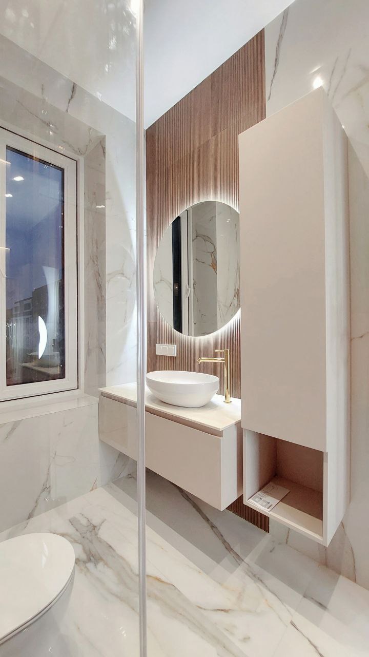 Nowoczesna łazienka z marmurowymi płytkami, okrągłym lustrem z podświetleniem LED, białą umywalką nablatową i złotą baterią, widoczna kabina prysznicowa ze szklanymi drzwiami.
