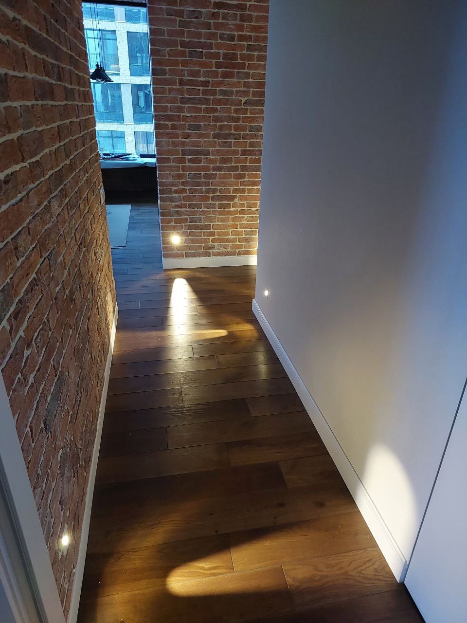 Wąski korytarz z podświetlanymi ceglanymi ścianami i drewnianą podłogą, widok z perspektywy wejścia, światło punktowe podkreśla fakturę cegły i drewna.