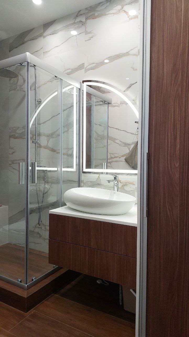 Nowoczesna łazienka z kabiną prysznicową, umywalką w kształcie łzy na szafce z forniru w odcieniach brązu oraz lustrem z podświetleniem LED w kształcie łuku.