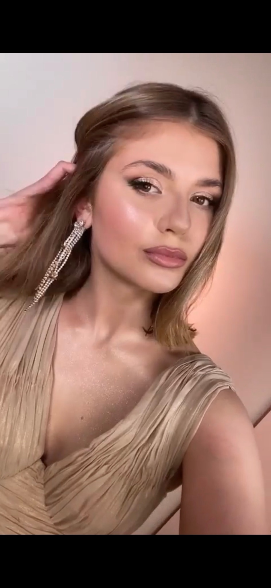 Portret kobiety z profesjonalnym, wieczorowym makijażem, długimi kolczykami z cyrkoniami i w drapowanej, złotej sukni. Widoczny subtelny tatuaż na szyi.