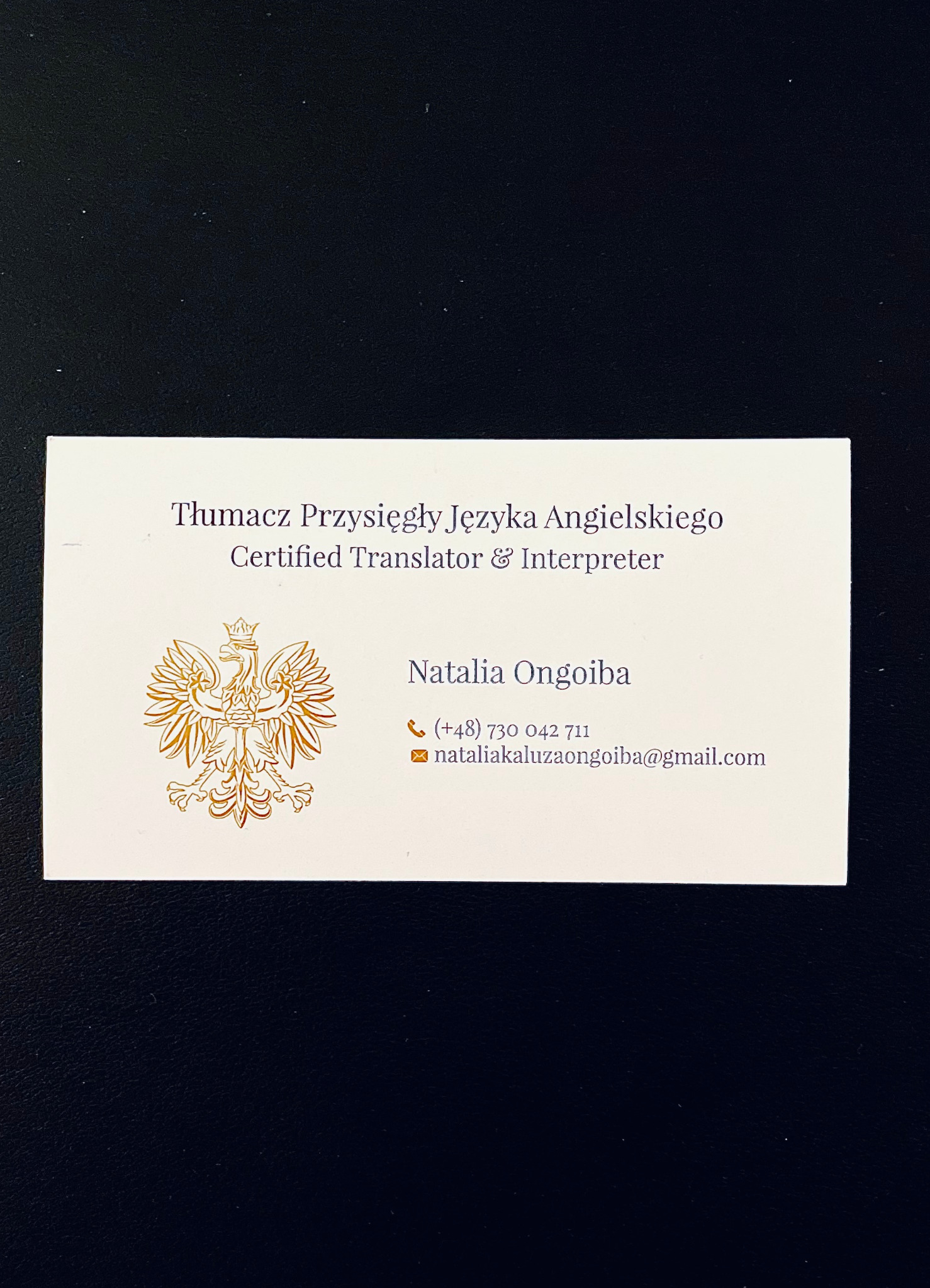 Wizytówka tłumacza przysięgłego języka angielskiego z godłem Polski, imieniem i nazwiskiem Natalia Ongoiba oraz danymi kontaktowymi, leżąca na ciemnym tle.