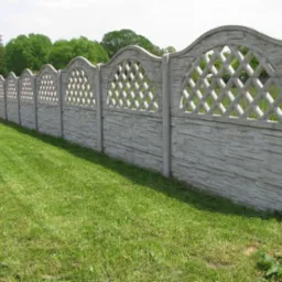 Długi, ozdobny płot z prefabrykowanych elementów betonowych z ażurowym wzorem rombów w górnej części, oddzielający trawnik od zalesionego terenu.