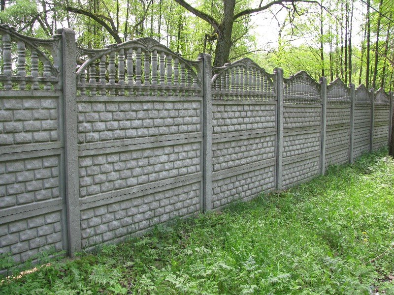 Długi, szary mur z prefabrykatów betonowych z dekoracyjnym, ażurowym wykończeniem u góry, imitujący cegłę, oddzielający teren od zieleni drzew i trawy.