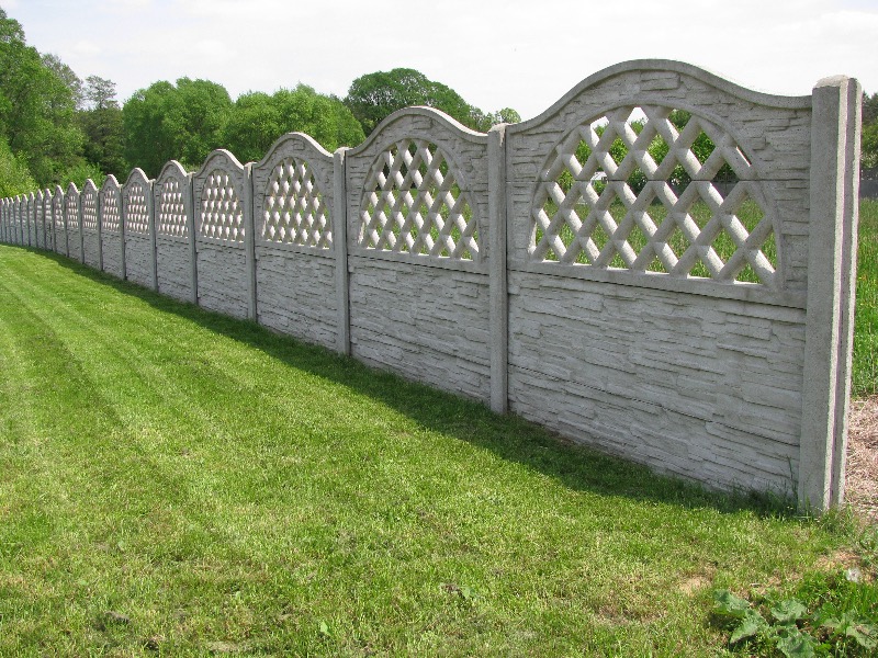 Długi, ozdobny płot z prefabrykowanych elementów betonowych z ażurowym wzorem rombów w górnej części, oddzielający trawnik od zalesionego terenu.