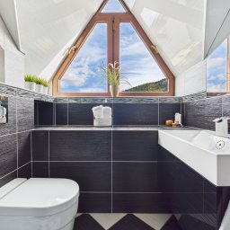 Agnieszka Pezda Nieruchomości - Nowoczesna łazienka na poddaszu z trójkątnym oknem, przez które widać las. Ciemne płytki kontrastują z białą armaturą i dodatkami. Nietypowy haczyk na ręcznik w kształcie stopy.