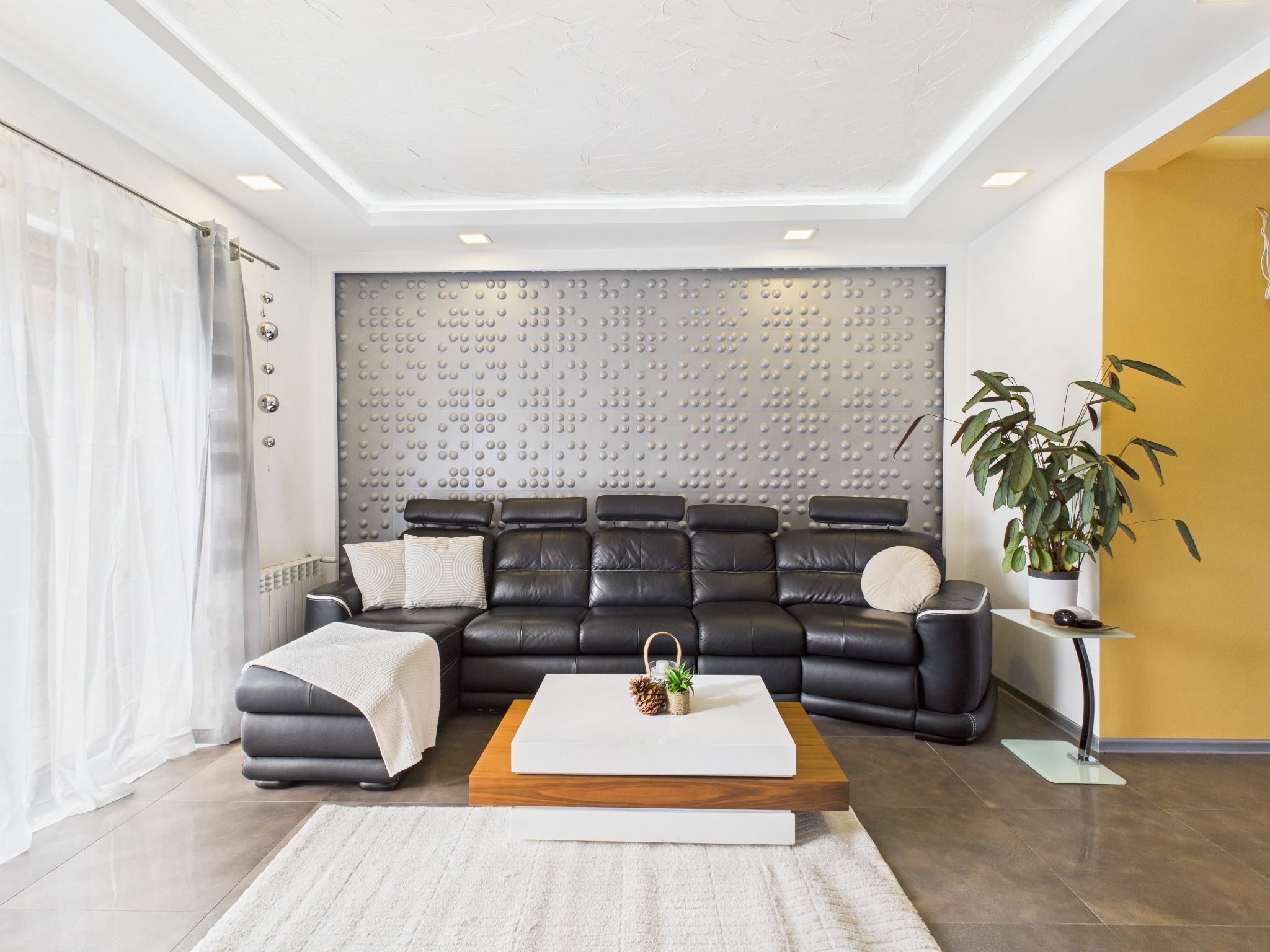 Nowoczesny salon z czarną skórzaną sofą, białym dywanem i ścianą z dekoracyjnymi panelami 3D. Roślina doniczkowa i stolik kawowy dopełniają aranżację.