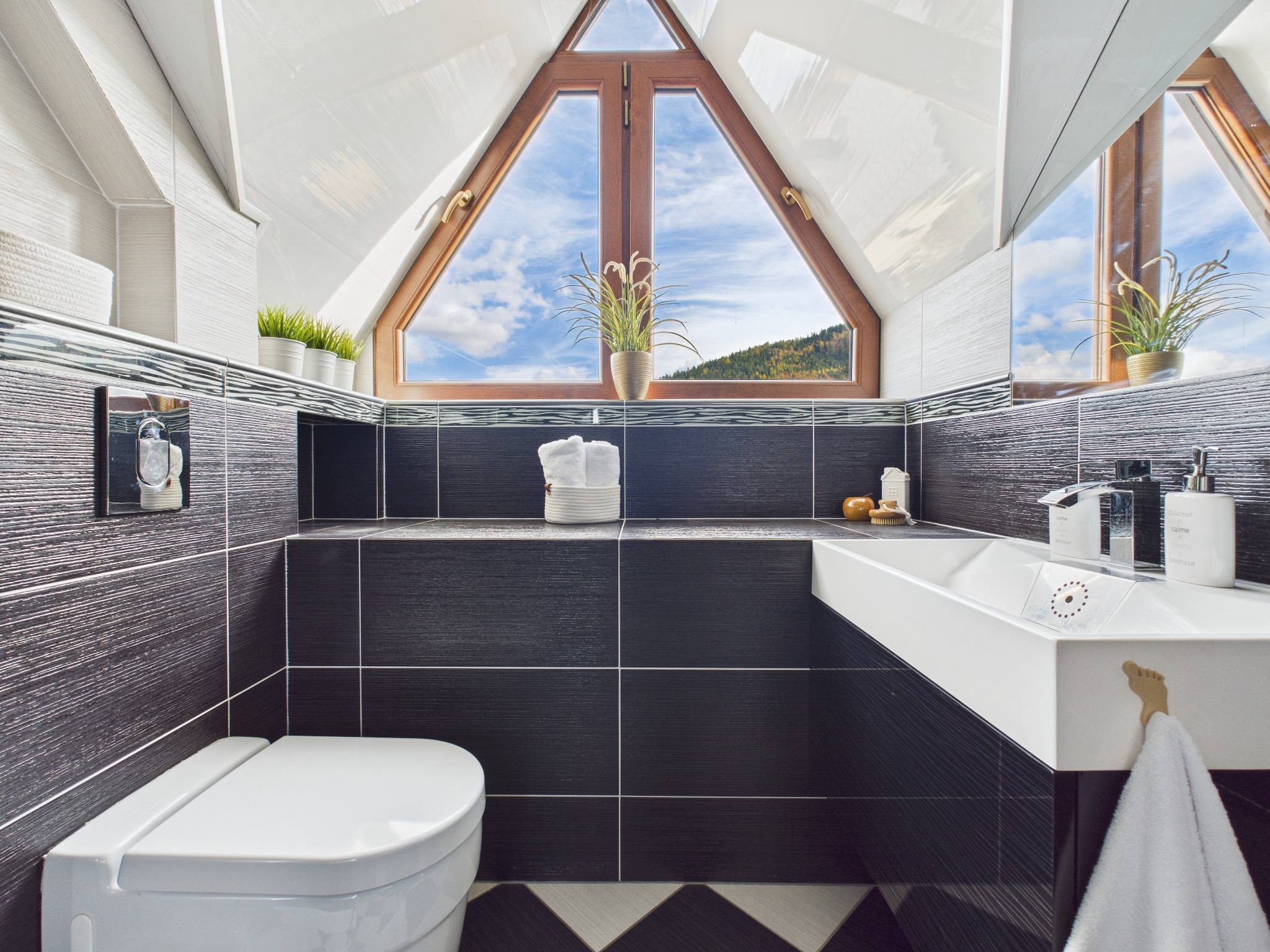 Nowoczesna łazienka na poddaszu z trójkątnym oknem, przez które widać las. Ciemne płytki kontrastują z białą armaturą i dodatkami. Nietypowy haczyk na ręcznik w kształcie stopy.