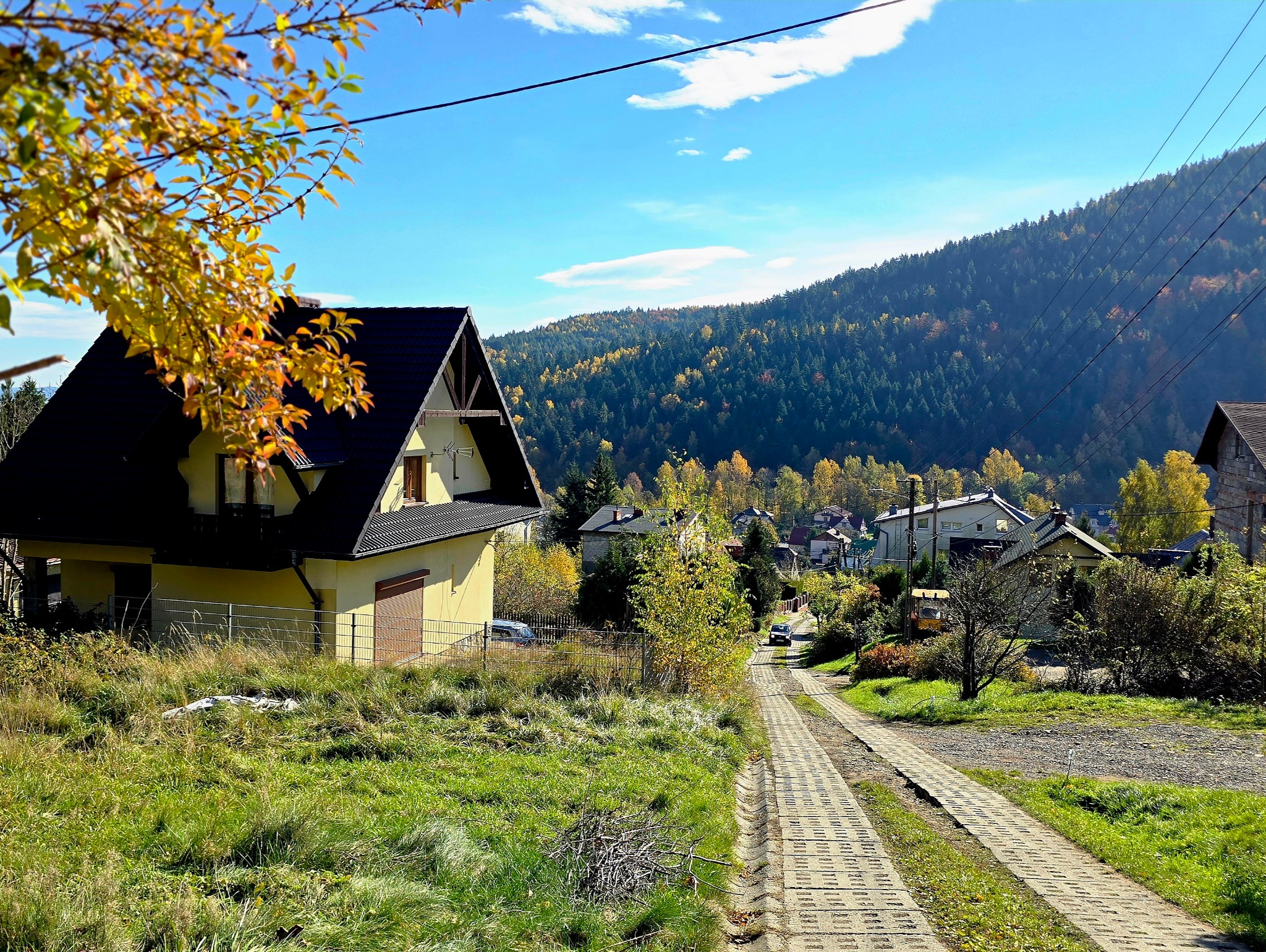 Żółty dom z czarnym dachem w jesiennej scenerii górskiej. Ujęcie z dołu, widoczna droga i zieleń. Słoneczny dzień, błękitne niebo.